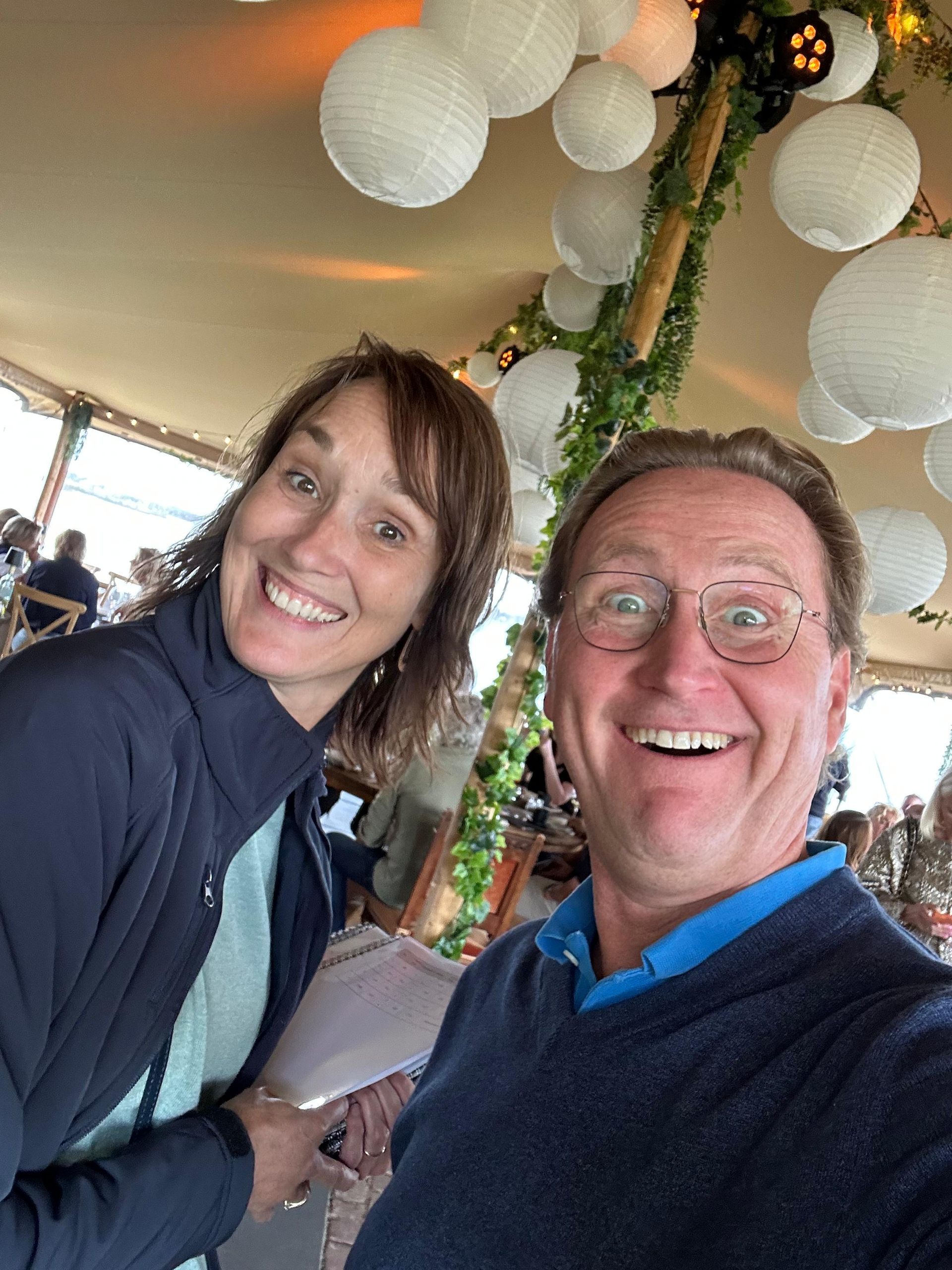 Een lachend stel maakt een selfie in een versierde tent, met witte lantaarns erboven.