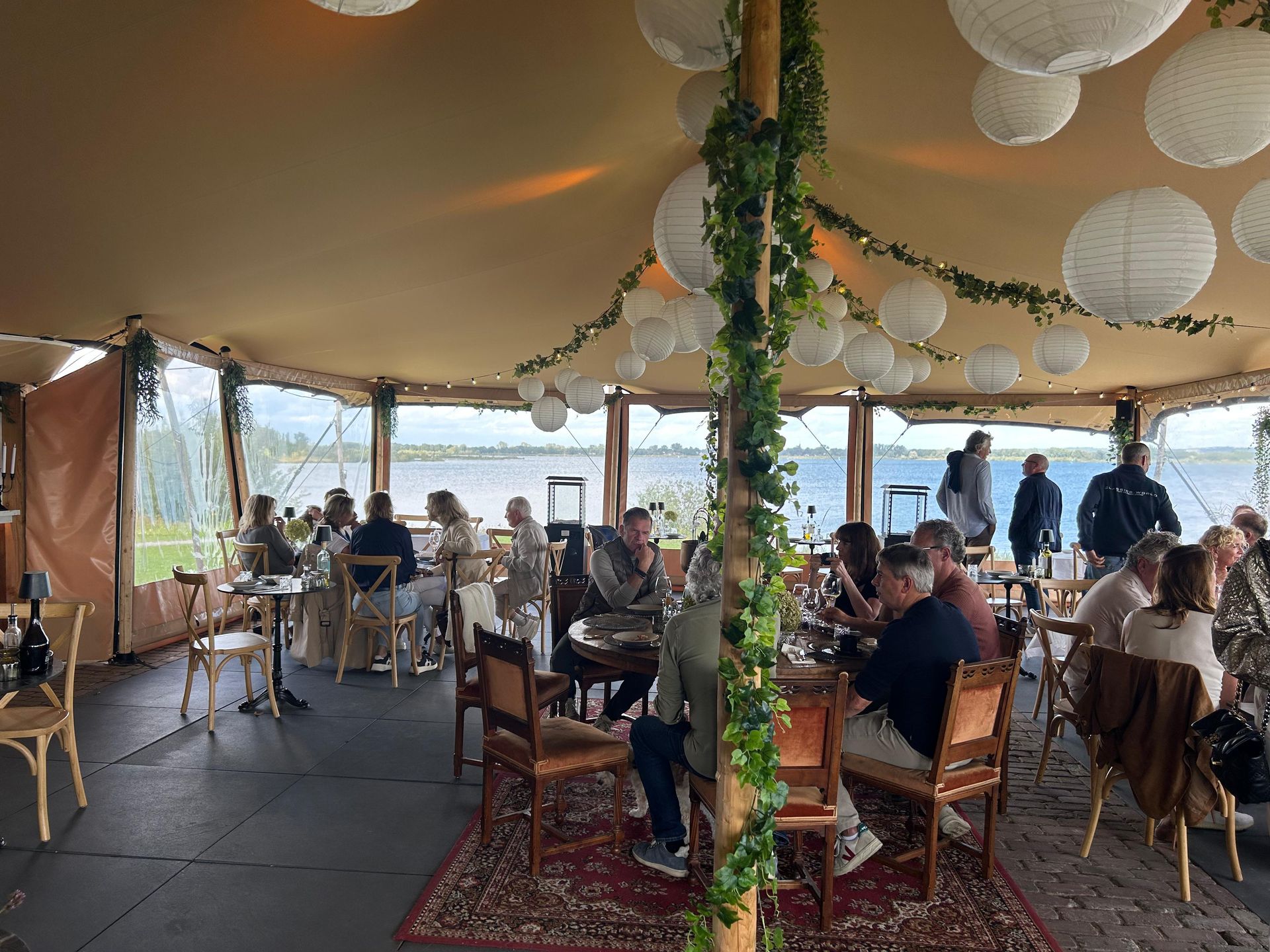 Mensen zitten aan tafels in een tent met uitzicht op het water. Witte papieren lantaarns en groen hangen aan het plafond.