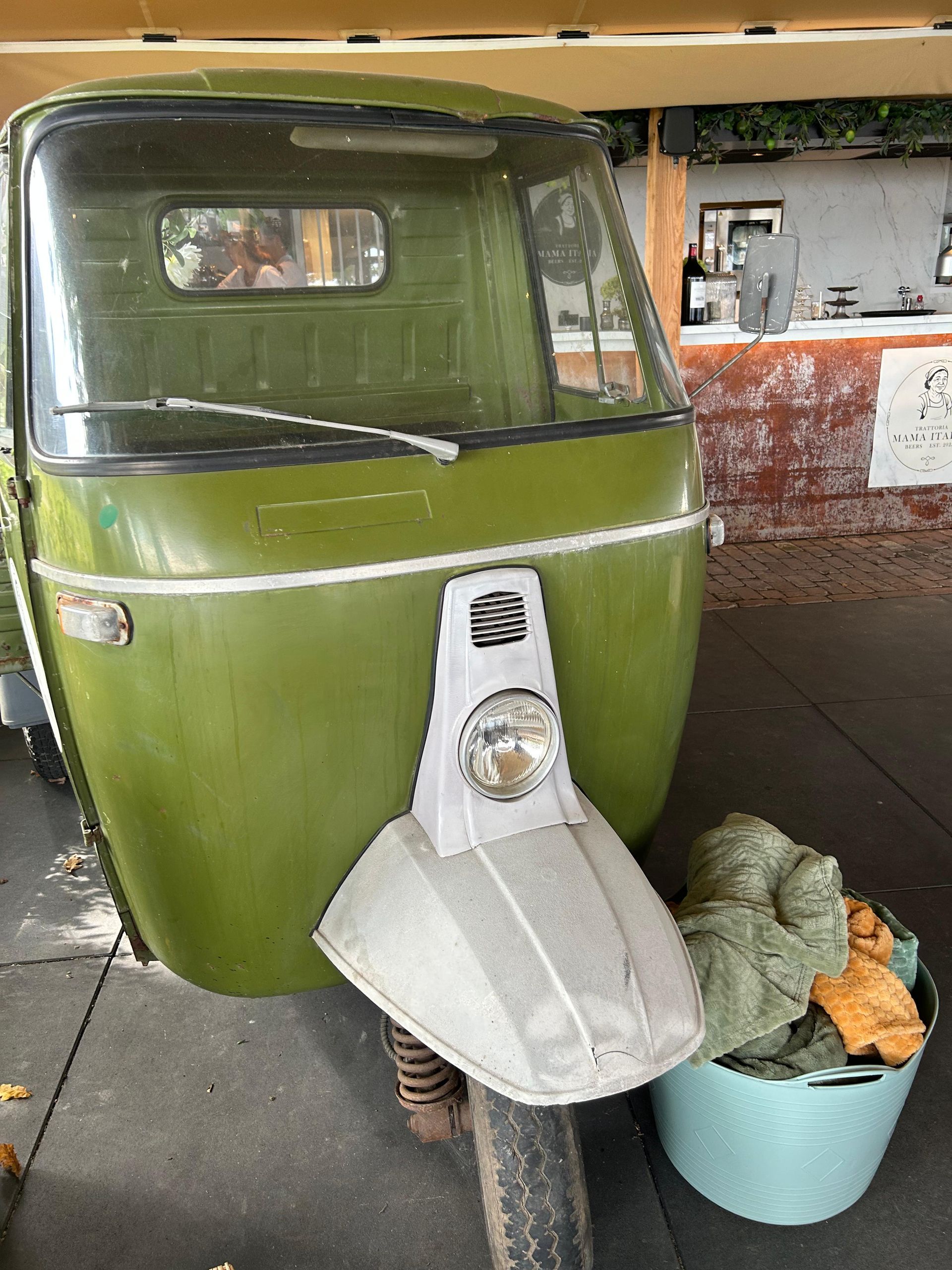 Groen-wit driewielig voertuig met een grote koplamp. Een turquoise emmer met doek ervoor.
