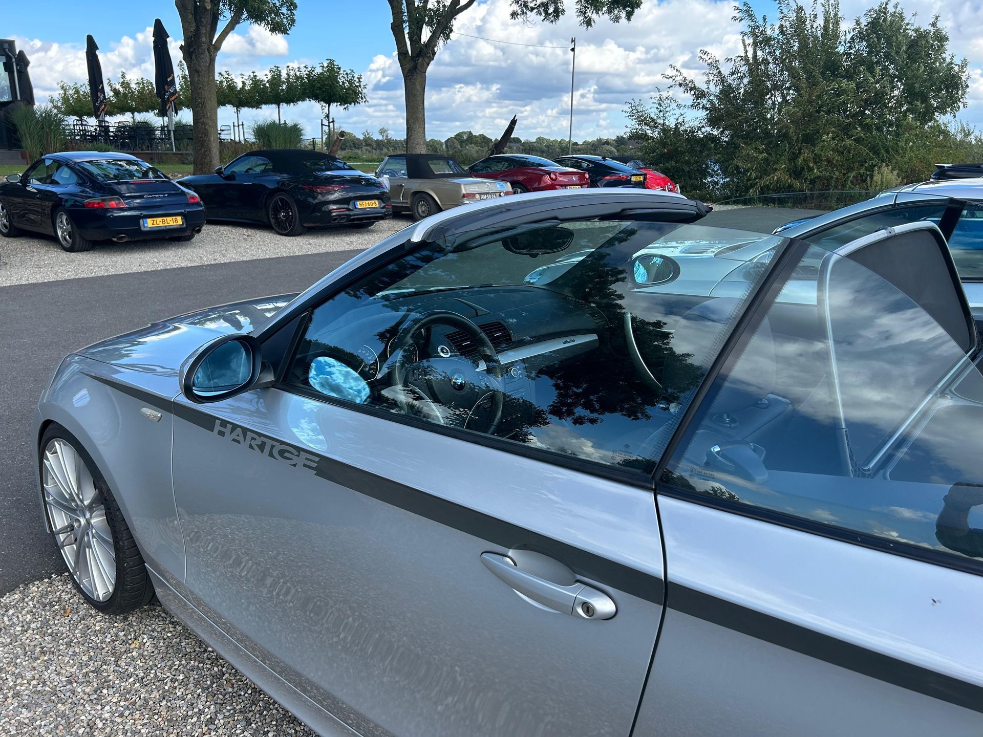 Een zilverkleurige cabriolet staat geparkeerd tussen andere auto's, met een blauwe lucht en bomen op de achtergrond.