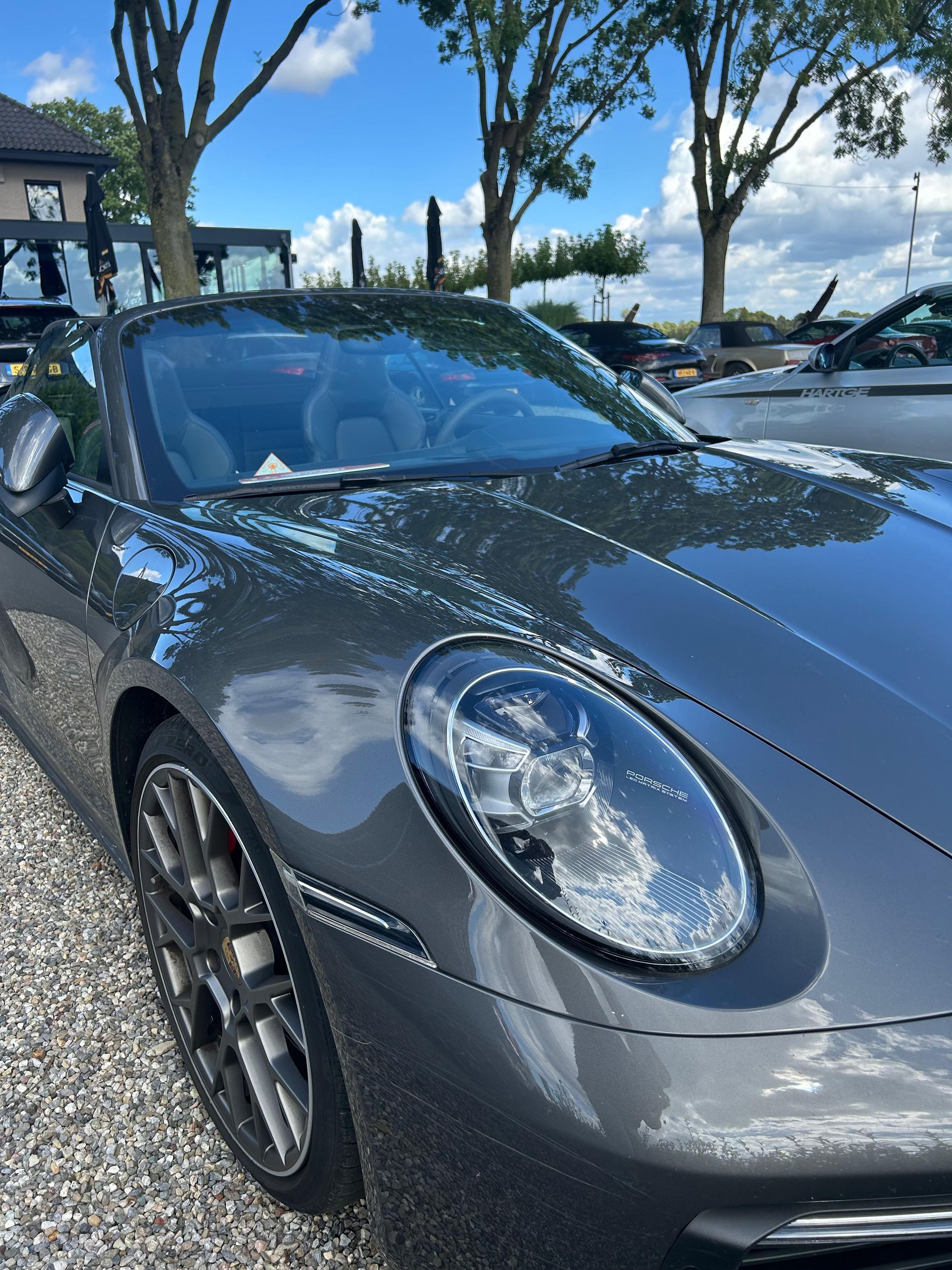 Een grijze Porsche cabriolet staat buiten geparkeerd onder een blauwe hemel, waar het zonlicht op weerkaatst.