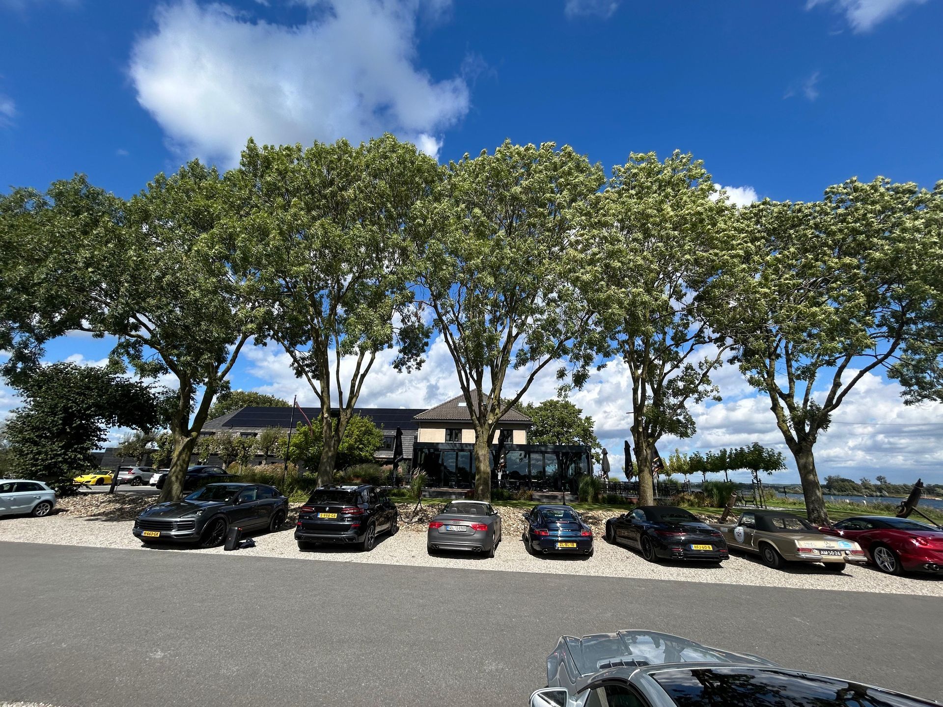 Auto's geparkeerd voor een gebouw, omzoomd door bomen, onder een blauwe hemel met wolken.