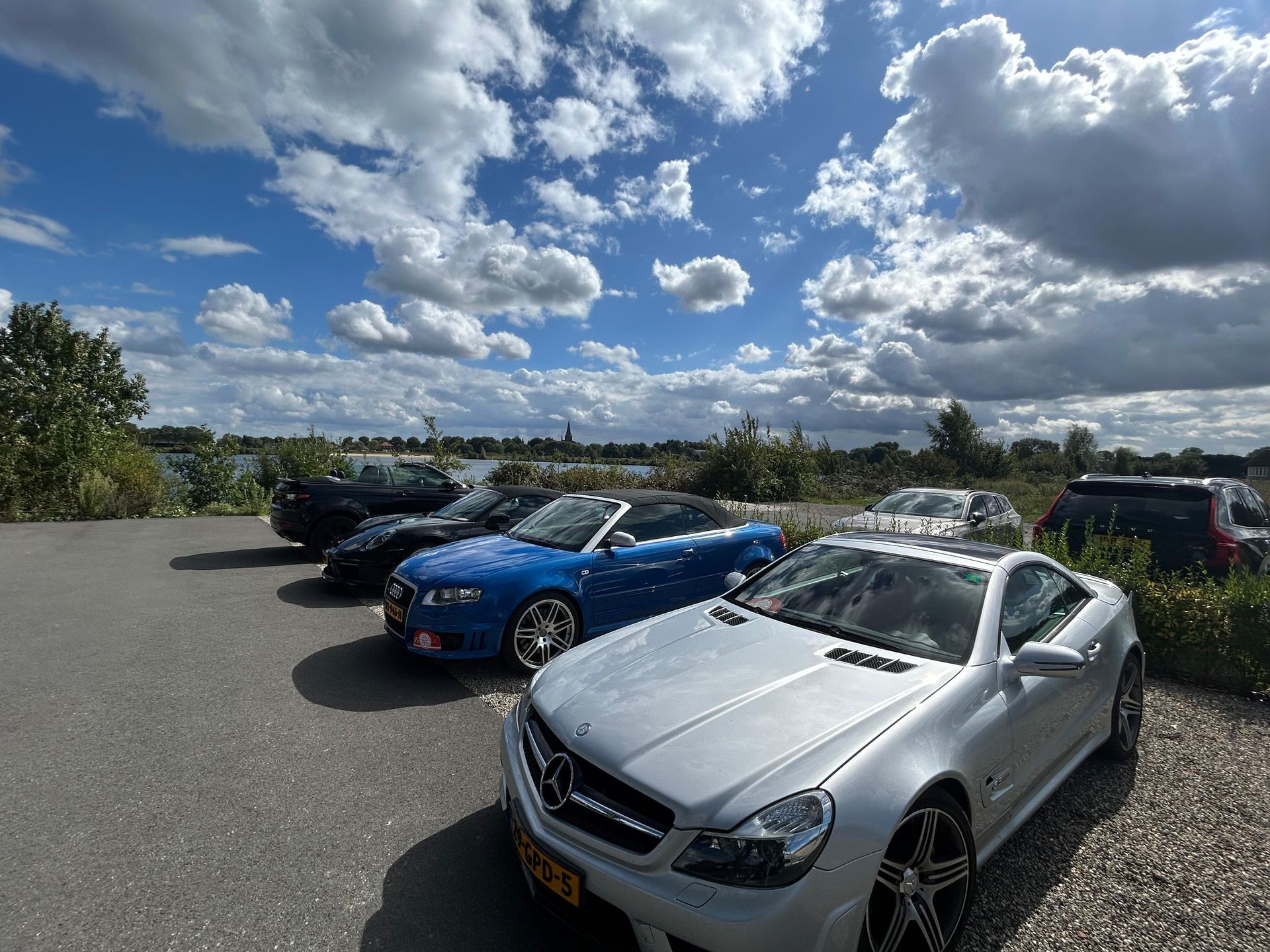 Auto's geparkeerd op een verhard terrein met een bewolkte blauwe lucht op de achtergrond. Zilverkleurige, blauwe en zwarte voertuigen.