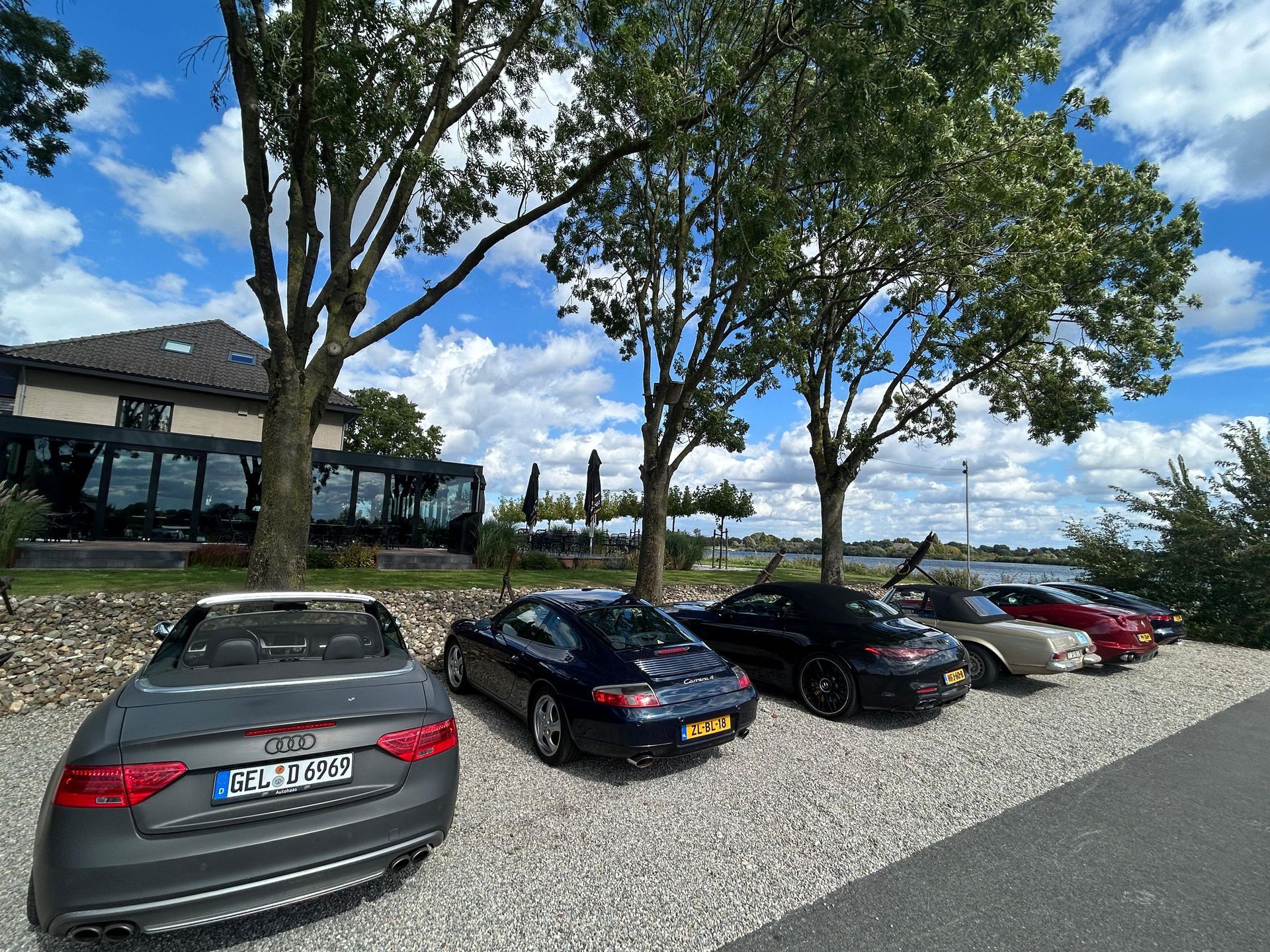 Auto's geparkeerd bij een gebouw met bomen onder een blauwe hemel. Een Audi cabriolet rijdt voorop.