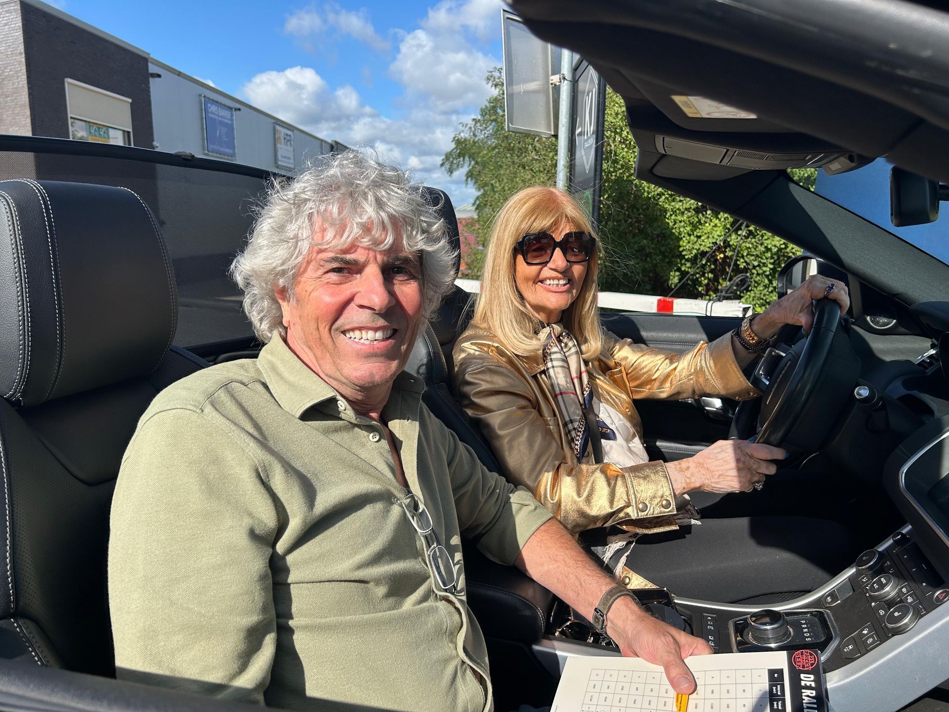 Twee mensen in een cabriolet, de vrouw bestuurt, beiden lachend. Zonnige dag, grijsbehaarde man, blonde vrouw.