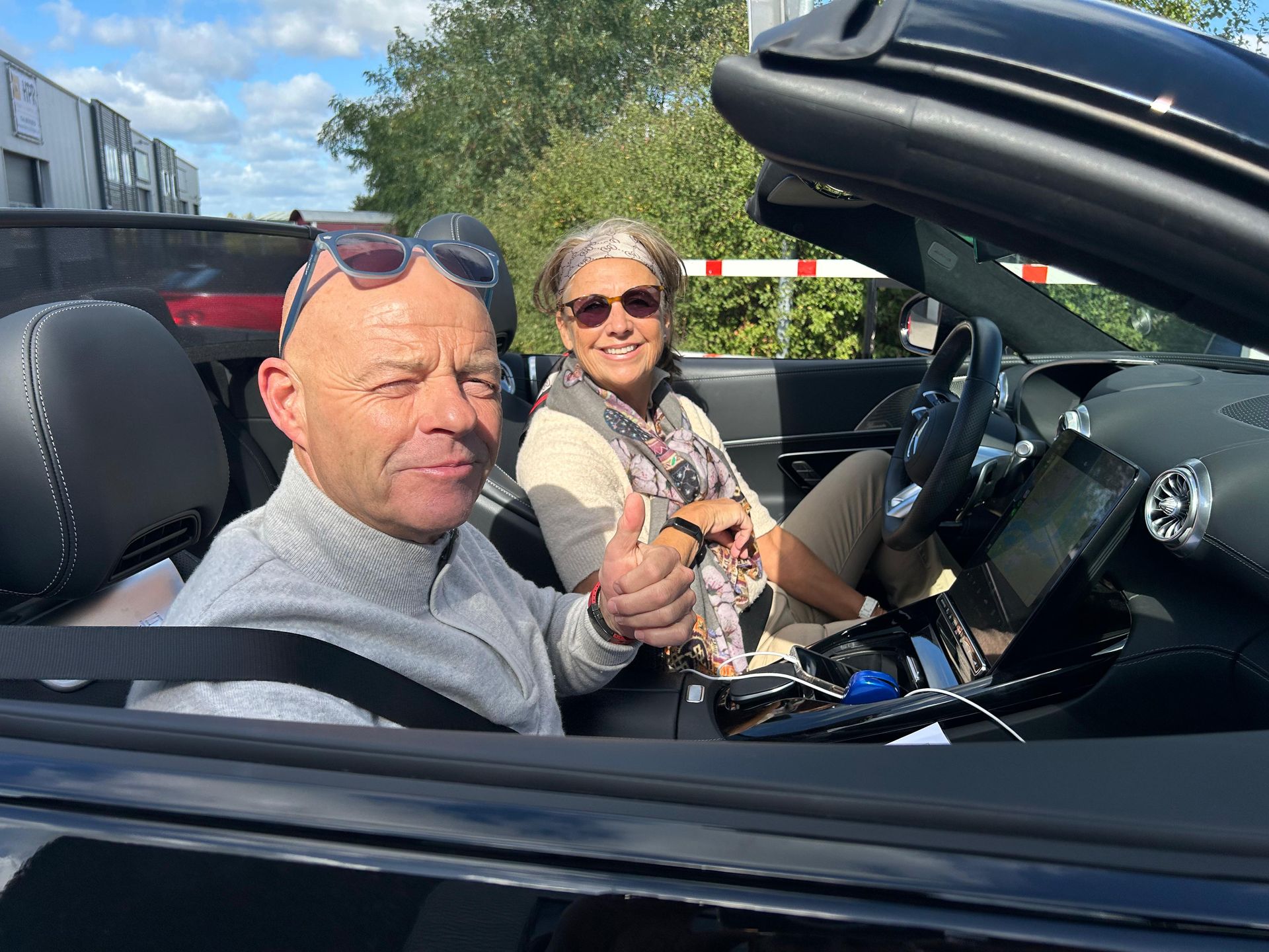 Een man steekt zijn duim omhoog, een vrouw glimlacht, zittend in een zwarte cabriolet, op een zonnige dag.