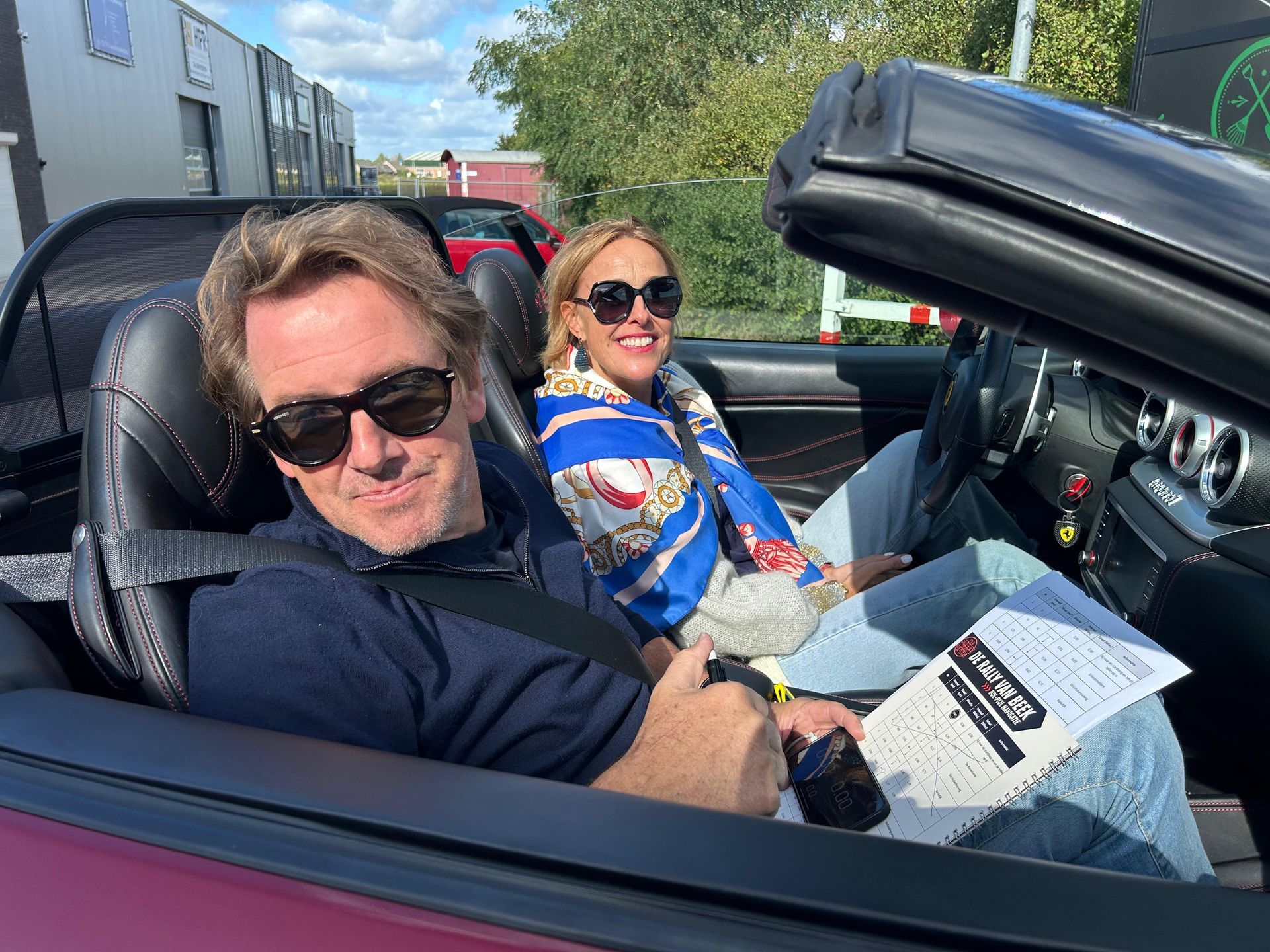 Een stel in een rode cabriolet, lachend. De man met zonnebril heeft een telefoon en papieren in zijn handen. De vrouw draagt een blauwe sjaal.