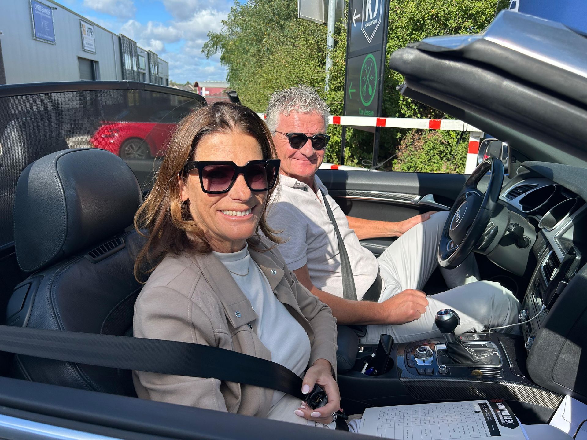 Een vrouw en een man met een zonnebril zitten lachend in een cabriolet. Buiten, zonnig weer.