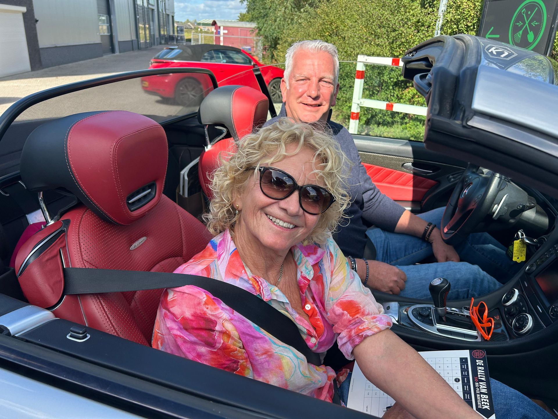 Een stel in een zilverkleurige cabriolet met rood interieur. De vrouw lacht en draagt een zonnebril.