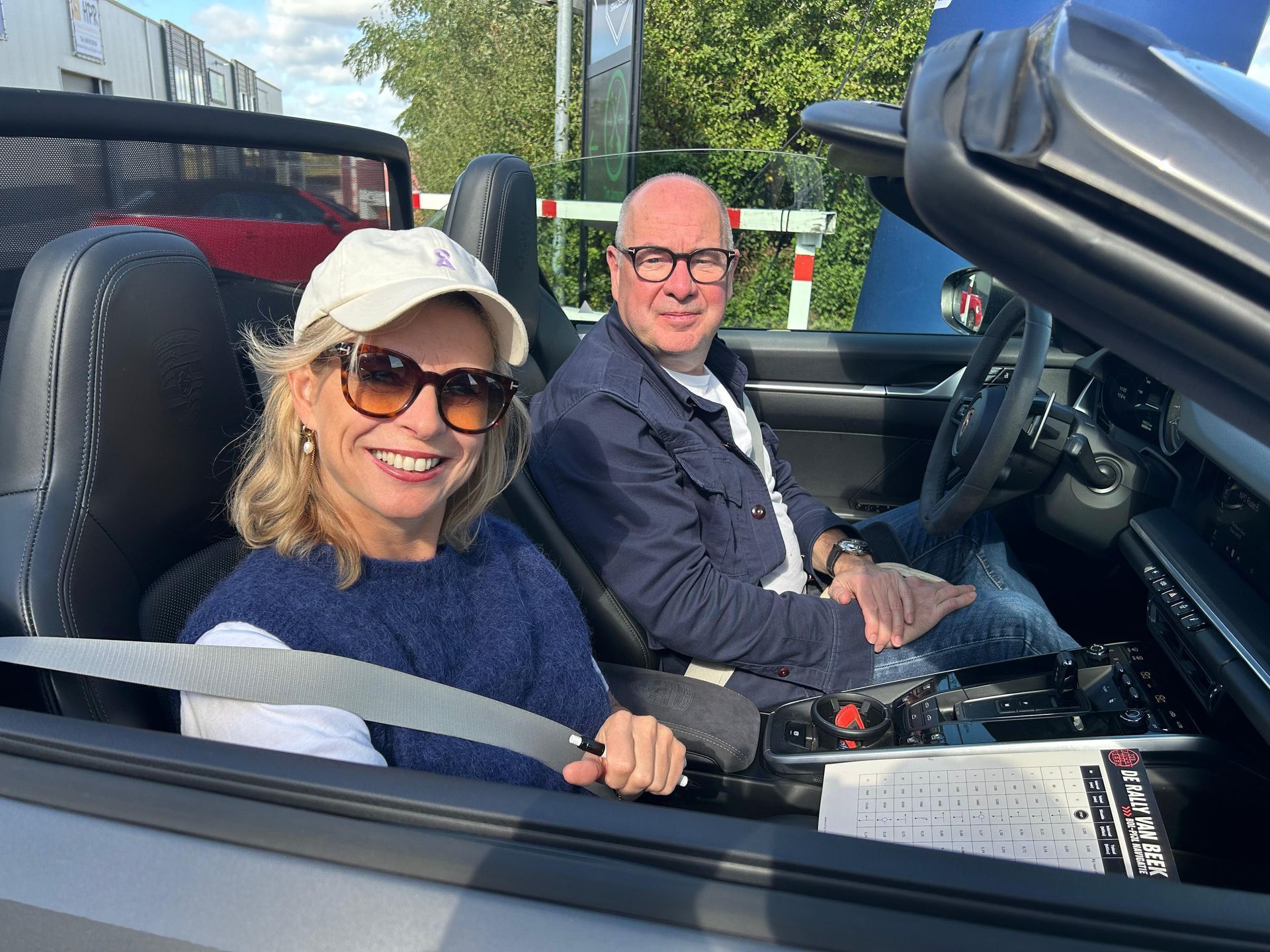 Vrouw en man glimlachen in een zilverkleurige cabriolet. De vrouw draagt een zonnebril en een pet. De man draagt een bril.