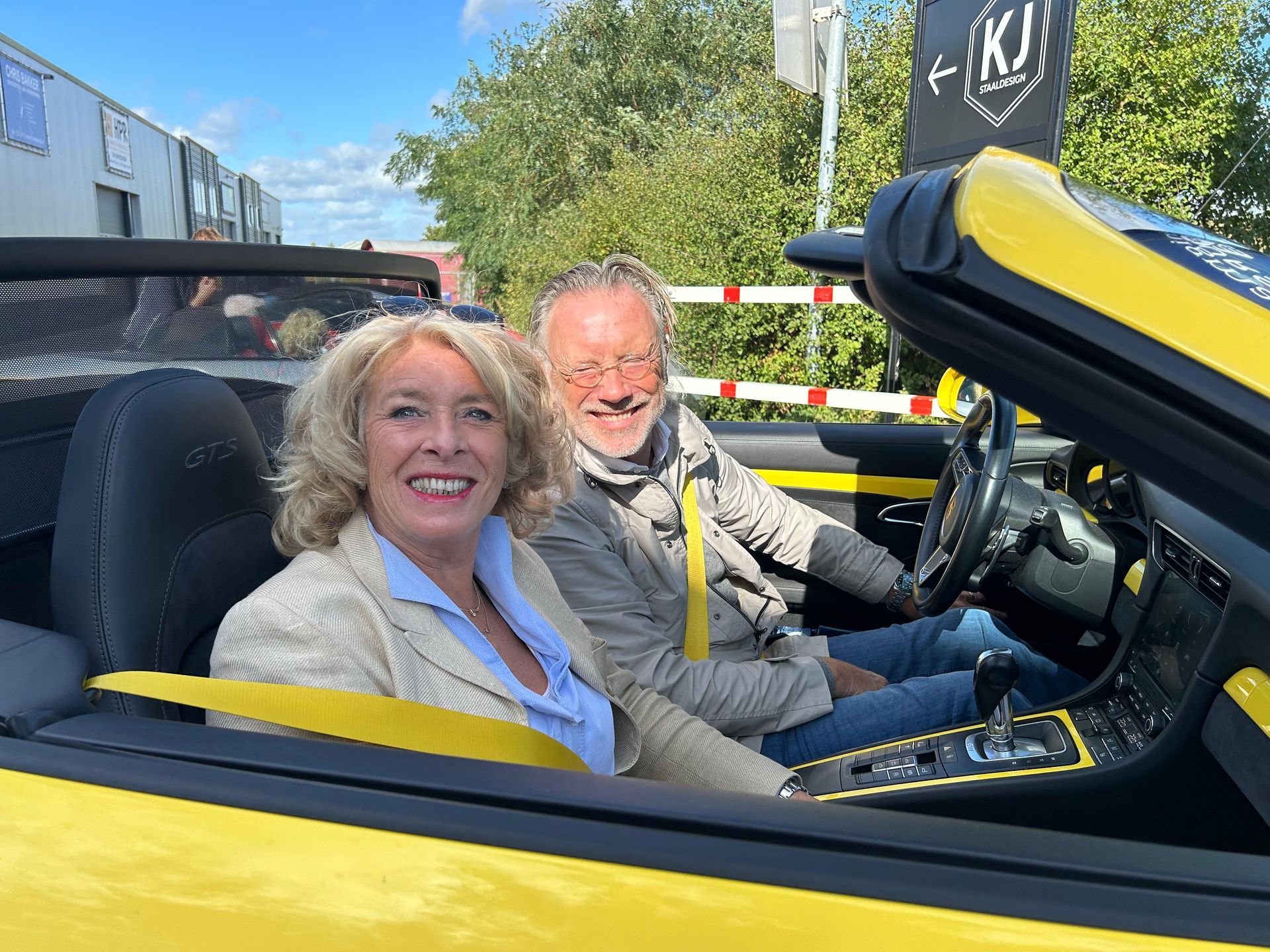 Een stel lacht vanuit een gele cabriolet. De vrouw draagt een dun jasje, de man een bril.