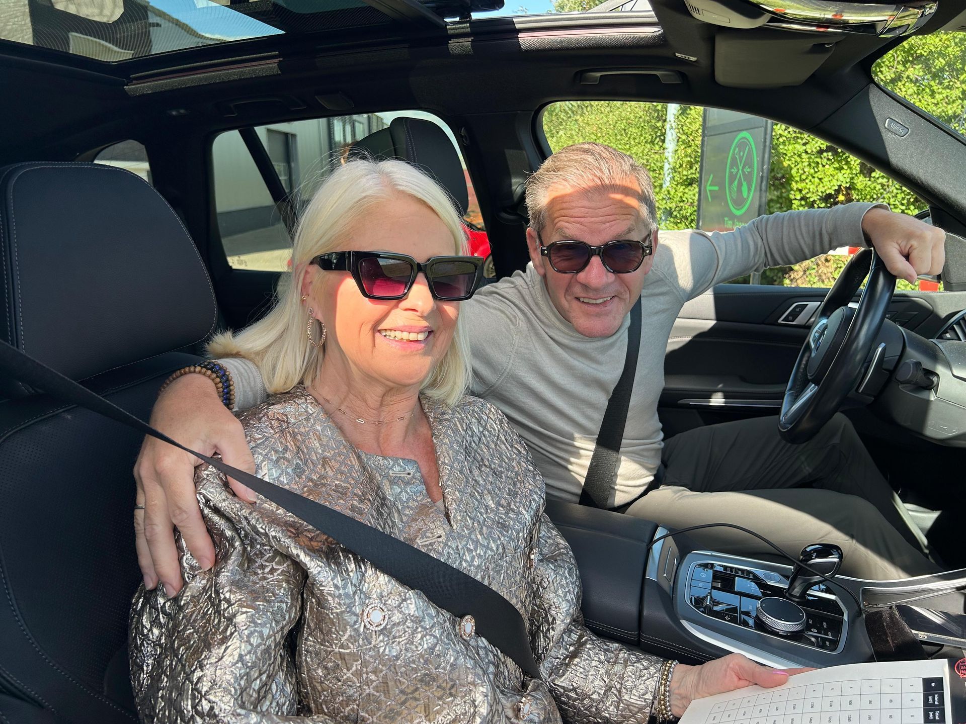 Twee mensen met zonnebrillen glimlachen in een auto; de man heeft zijn arm om de vrouw heen geslagen.