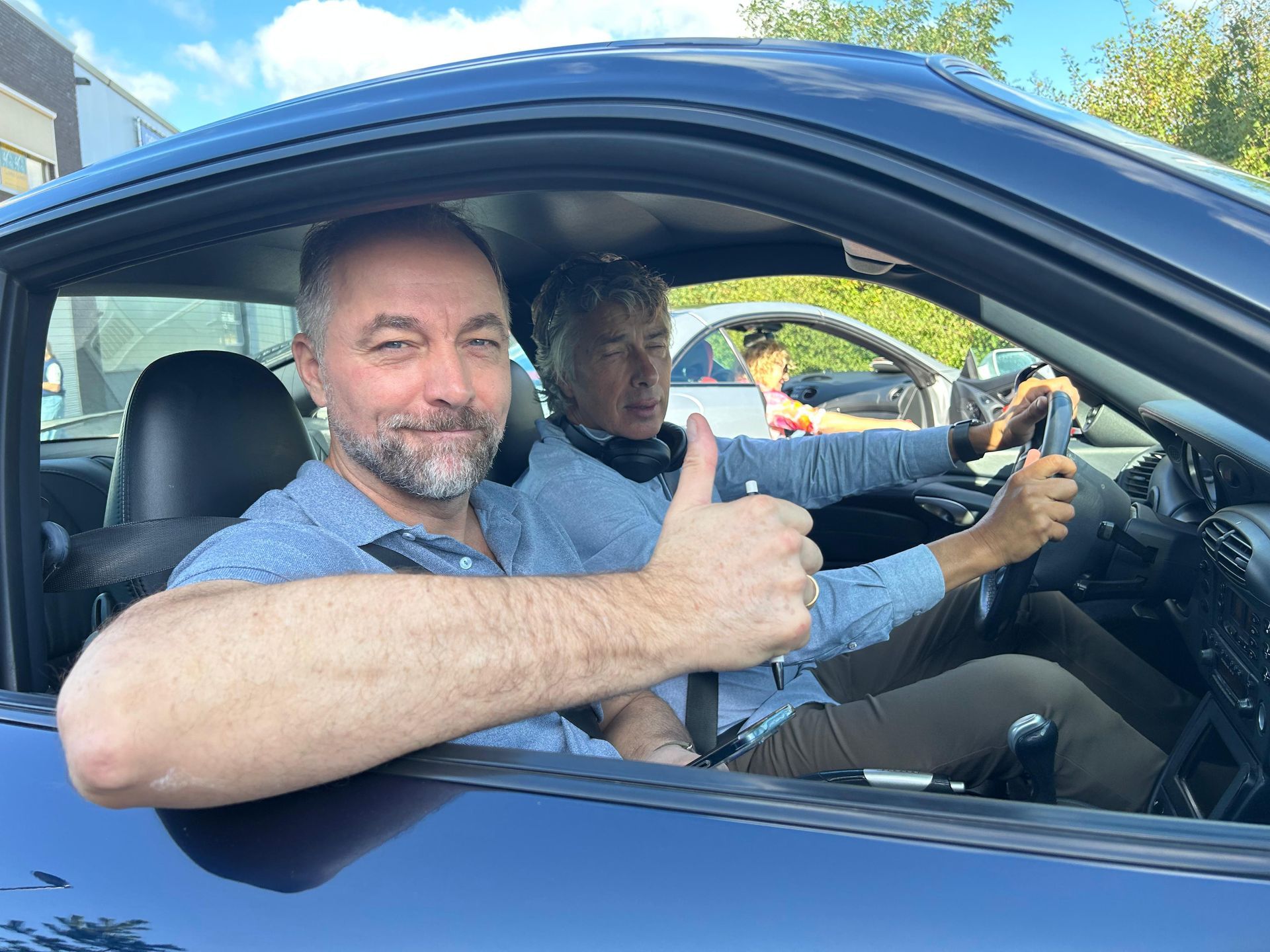 Twee mannen in een blauwe auto; een van hen steekt zijn duim omhoog. Een andere auto is zichtbaar op de achtergrond. Zonnige dag.
