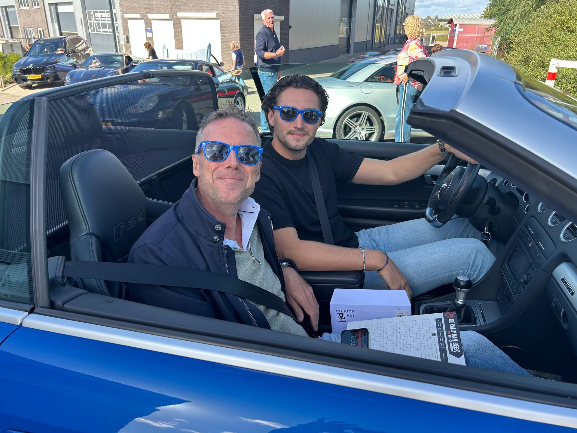 Twee mannen in een blauwe cabriolet, lachend en met zonnebrillen op, rijdend. Andere auto's op de achtergrond.