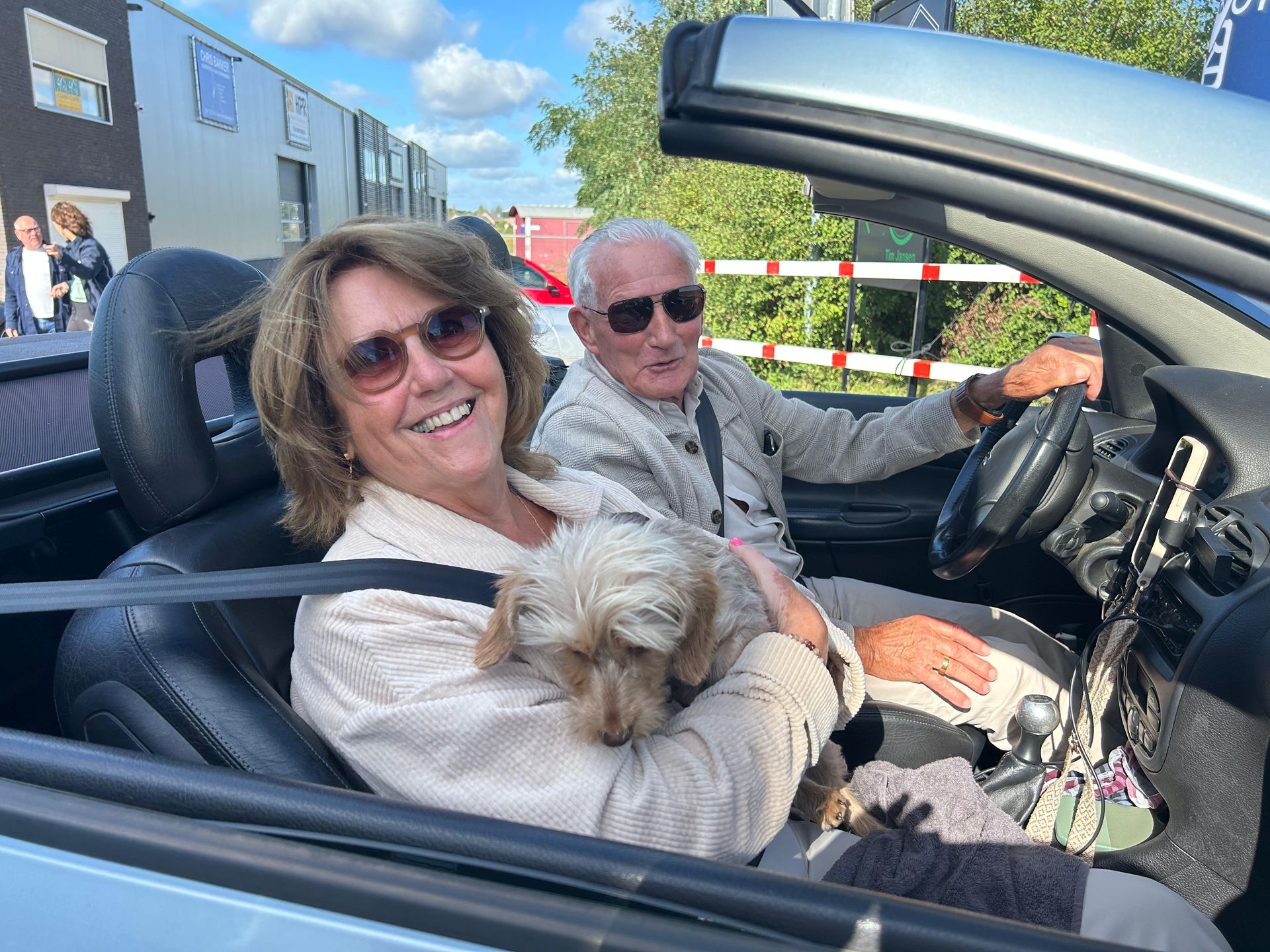 Paar in cabriolet met hond; vrouw lacht, man rijdt; zonnige dag.