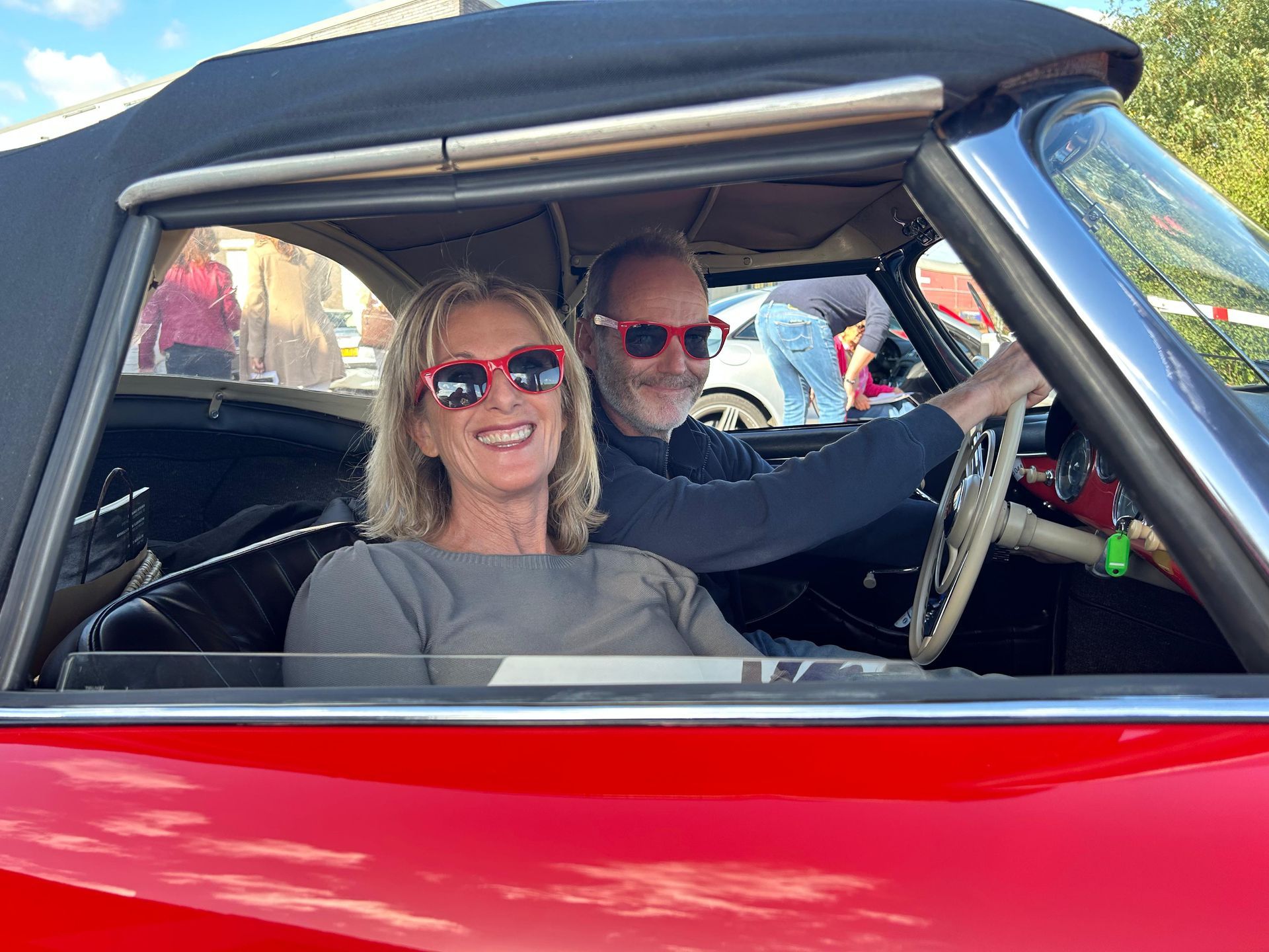 Een stel met rode zonnebrillen in een rode cabriolet. De vrouw lacht, de man rijdt, de ramen van de auto staan open.