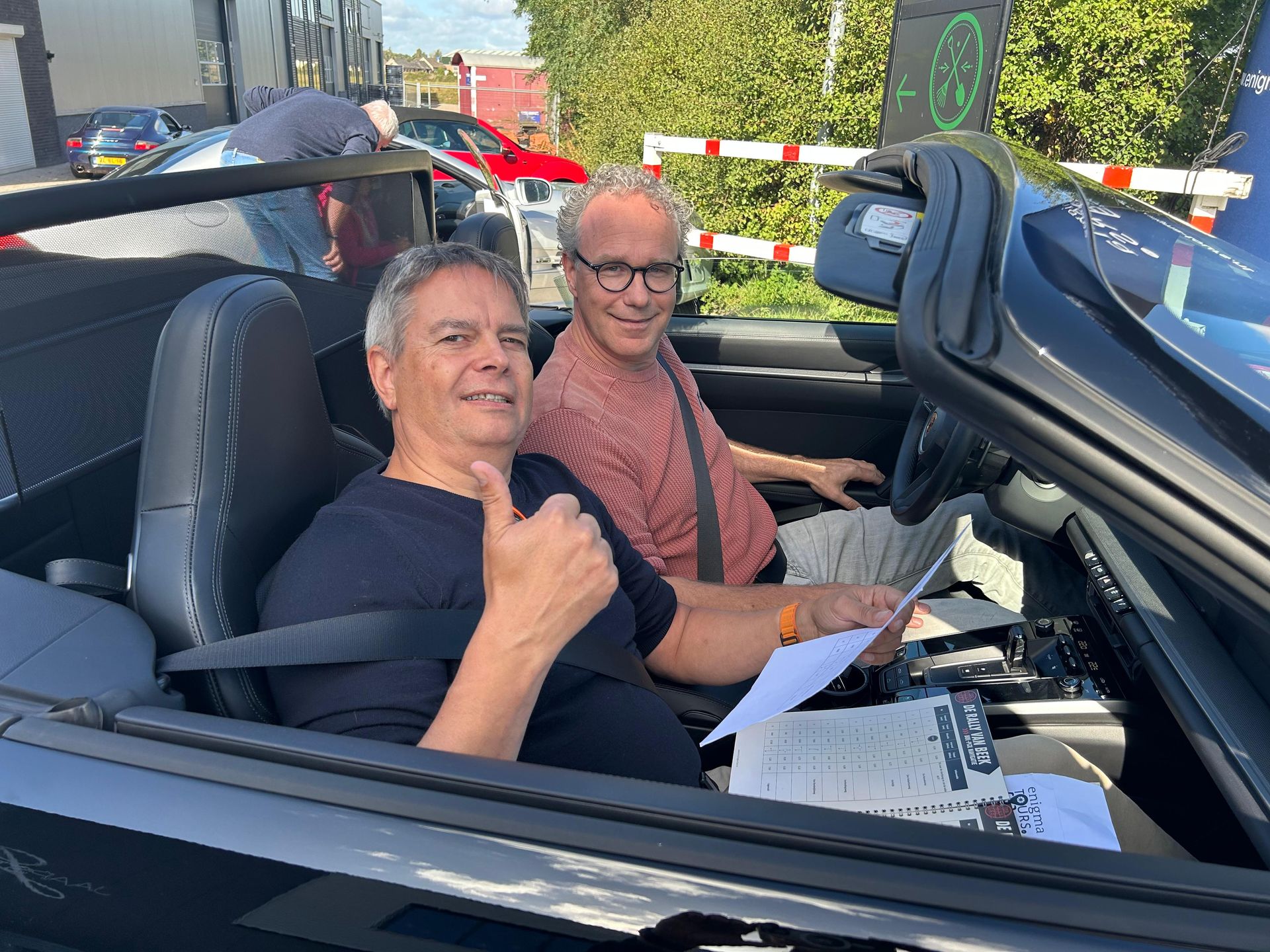 Twee mannen in een zwarte cabriolet; een van hen steekt zijn duim omhoog. Een heldere zonnige dag, een vangrail op de achtergrond.