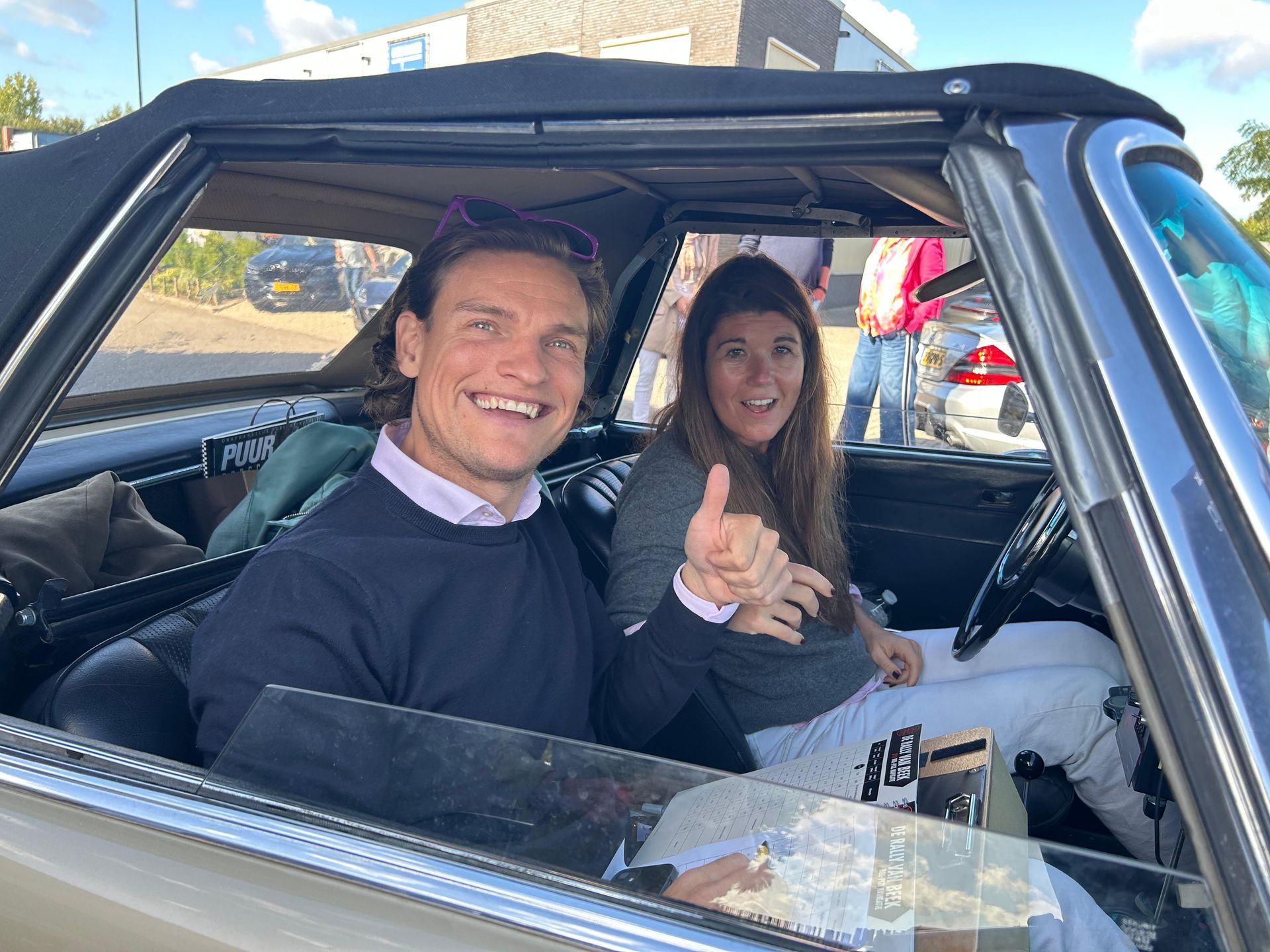 Twee lachende mensen in een cabriolet. De man bestuurt de auto, de vrouw steekt haar duim omhoog; beiden bevinden zich buiten.