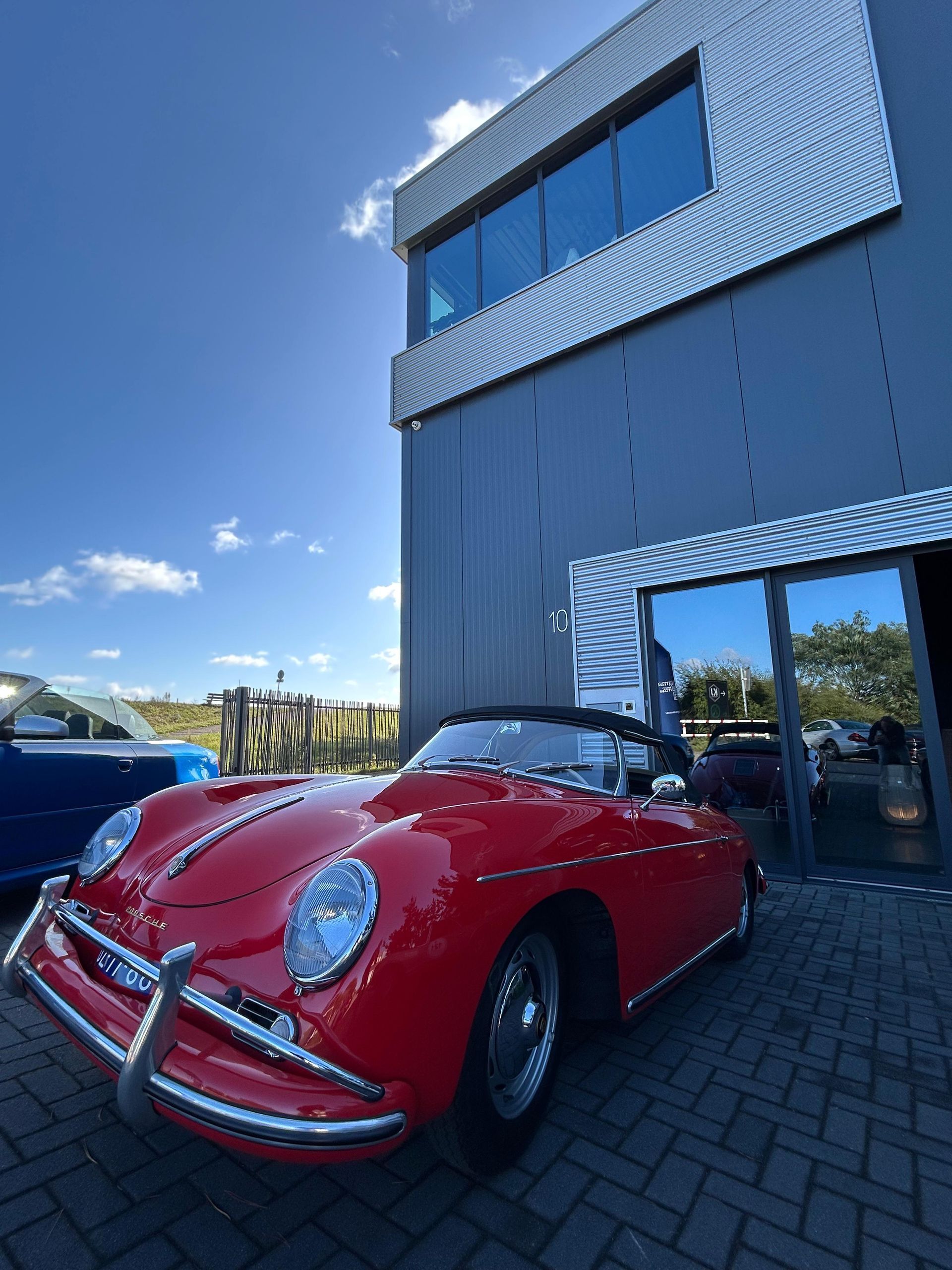 Een rode vintage Porsche cabriolet staat geparkeerd voor een modern gebouw op een zonnige dag.