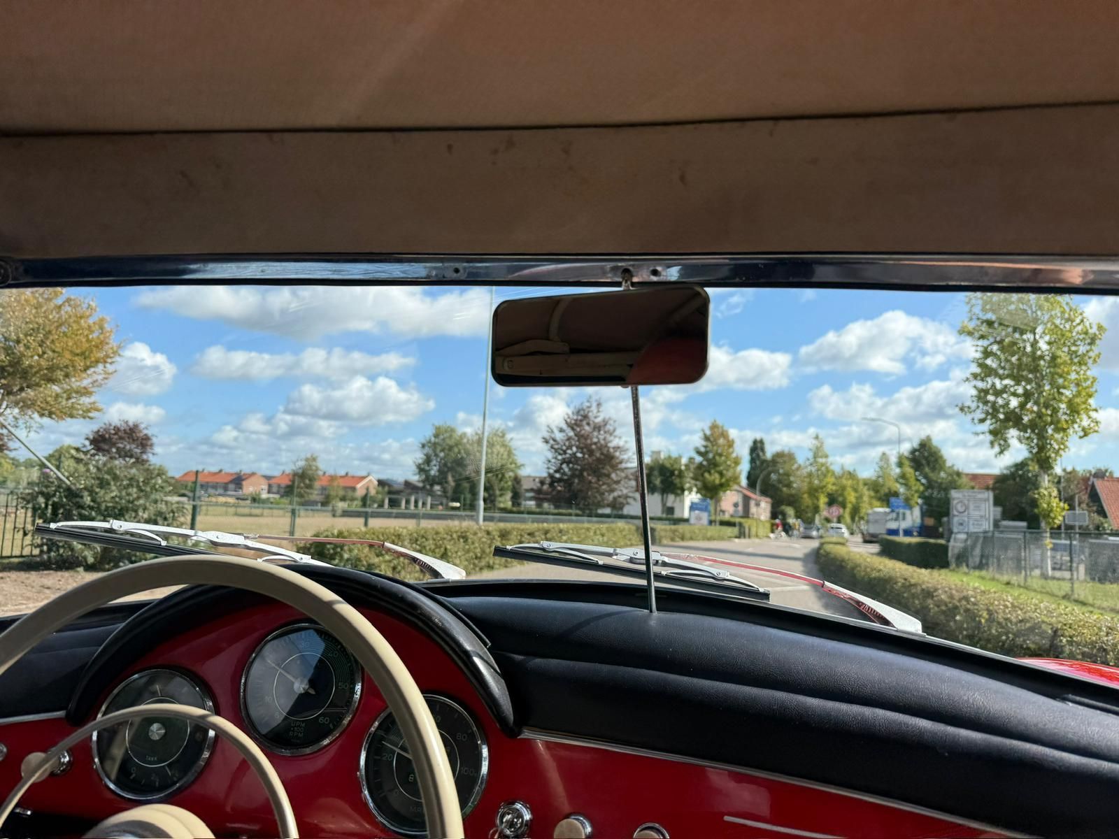 Uitzicht vanuit een klassieke rode auto. Het dashboard en het stuur zijn zichtbaar, met een straat en de lucht in de verte.
