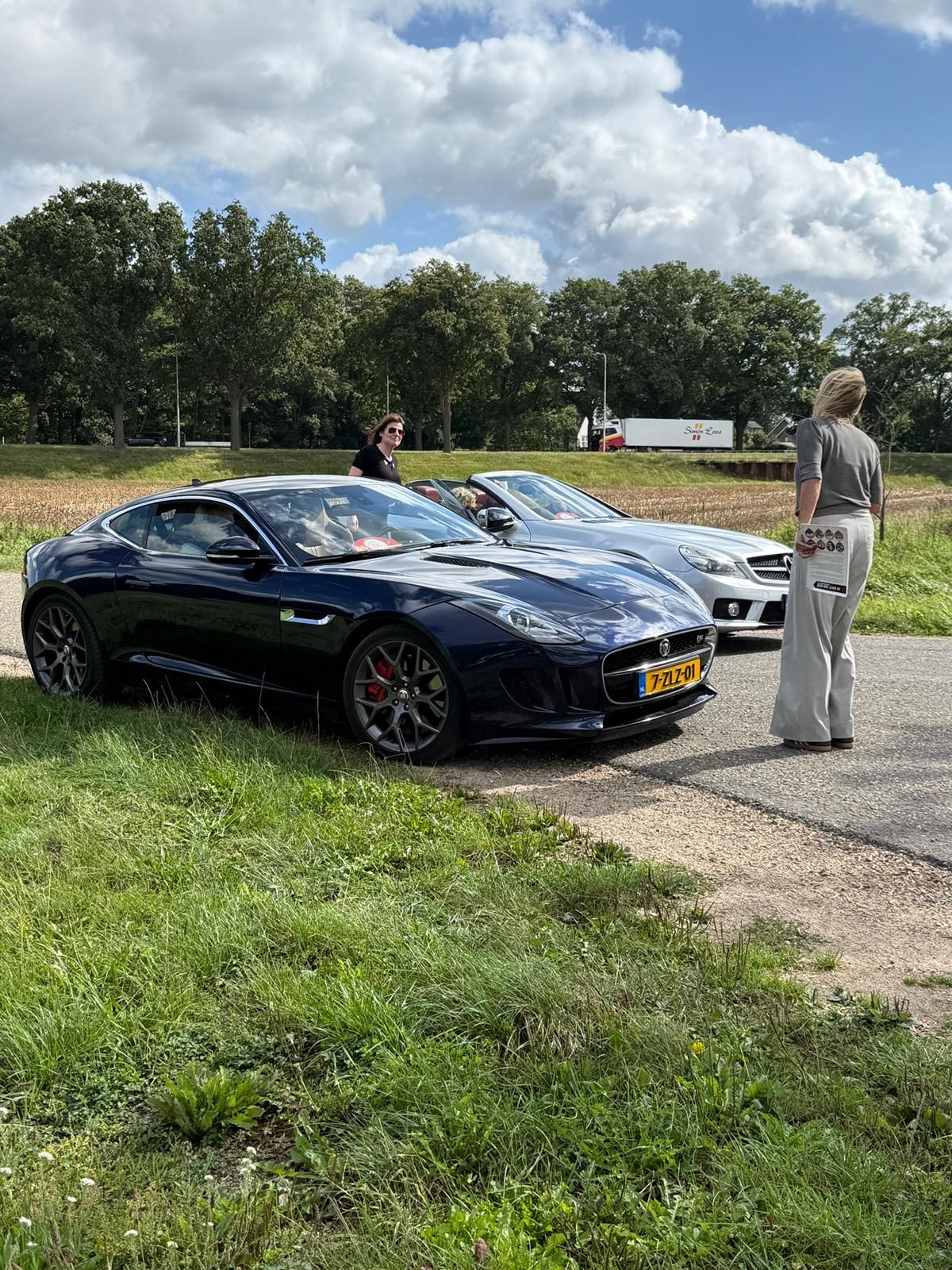 Een zwarte Jaguar sportwagen staat geparkeerd naast een zilverkleurige auto, met een persoon er vlakbij. Buiten op een zonnige dag.