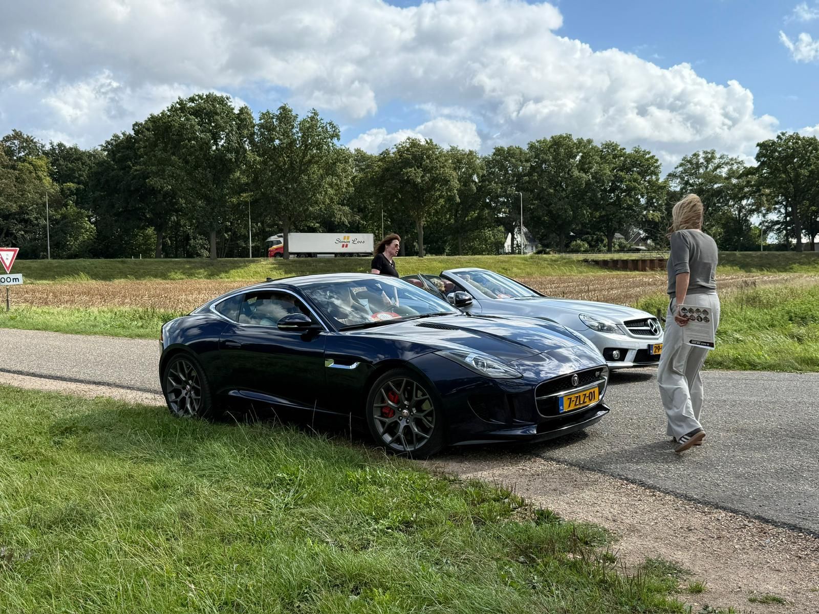 Twee auto's geparkeerd langs de weg. Een donkerblauwe Jaguar en een zilveren Mercedes, met twee mensen die er vlakbij staan.