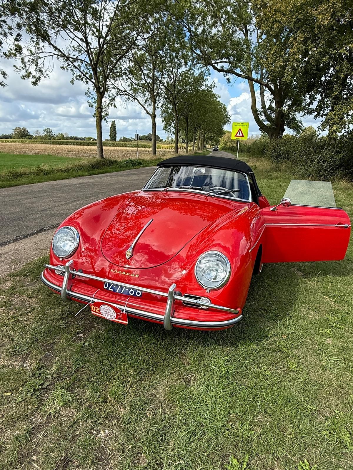 Een rode vintage Porsche cabriolet met open deur staat geparkeerd op het gras langs de weg.
