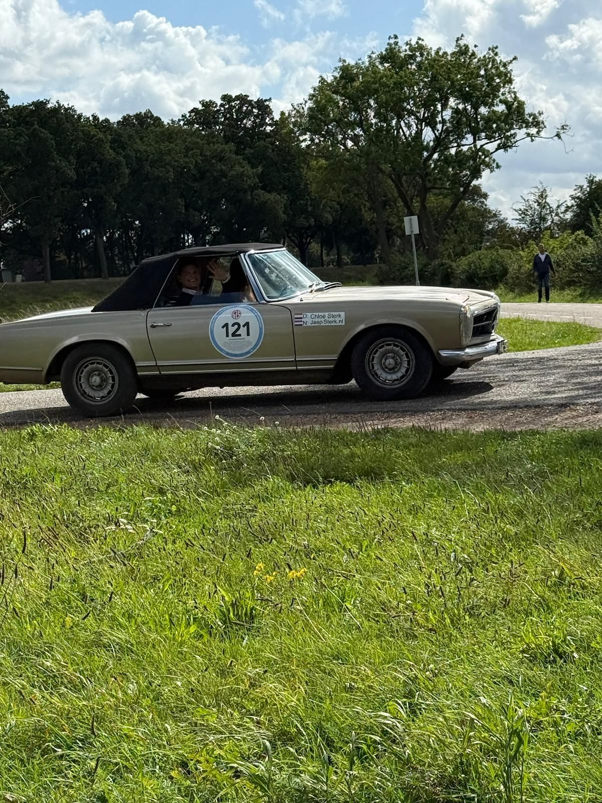 Een beige vintage cabriolet met twee passagiers op een weg, met bomen en gras op de achtergrond.