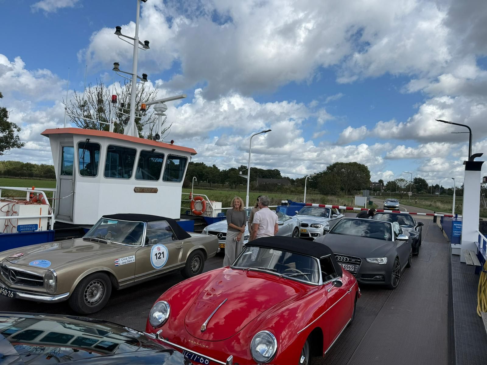 Klassieke auto's op een veerboot, een rode Porsche vooraan, andere auto's in een rij, passagiers in de buurt, bewolkte lucht.