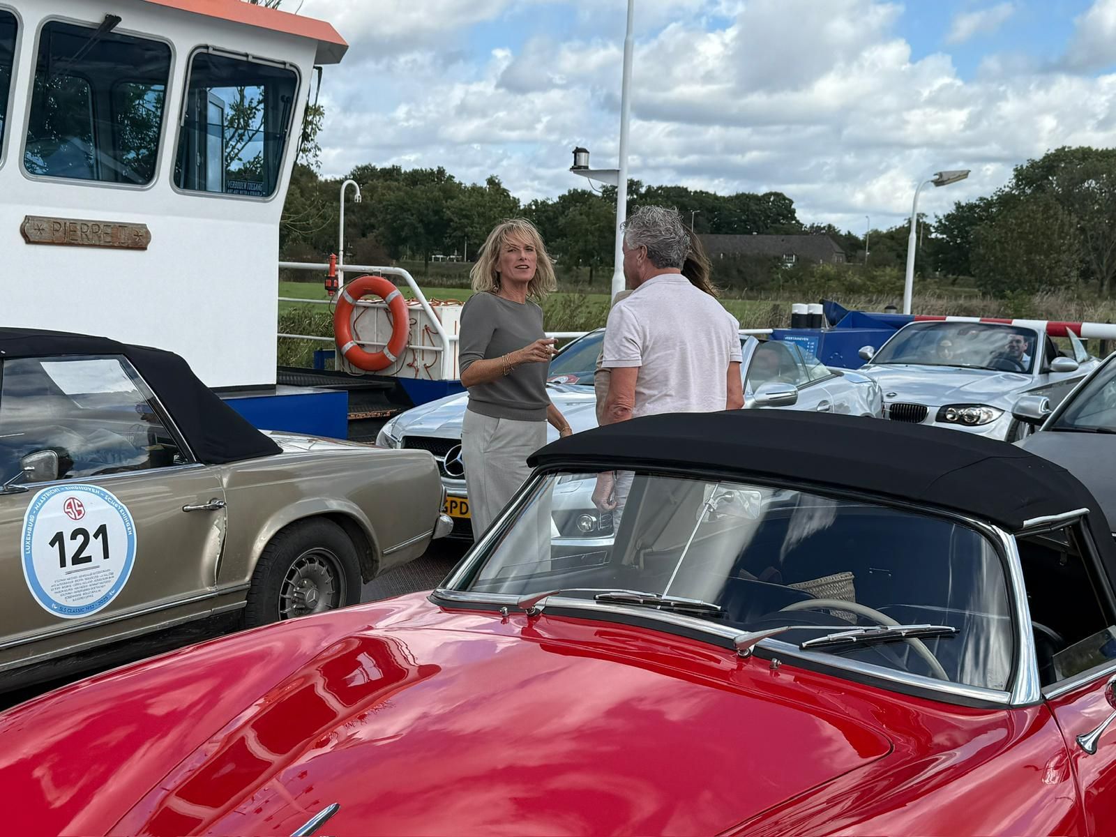 Een vrouw en een man voeren een gesprek in de buurt van klassieke cabriolets op een veerboot.