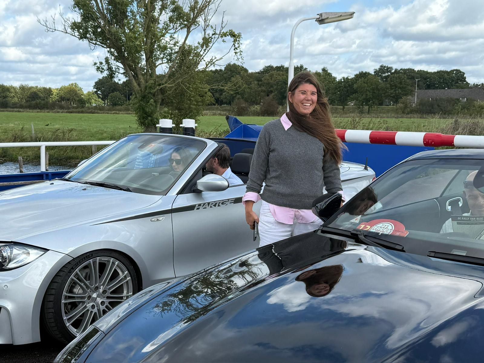 Een vrouw staat lachend tussen twee cabriolets. De auto's zijn zilver en zwart, en de omgeving lijkt een veerboot te zijn.