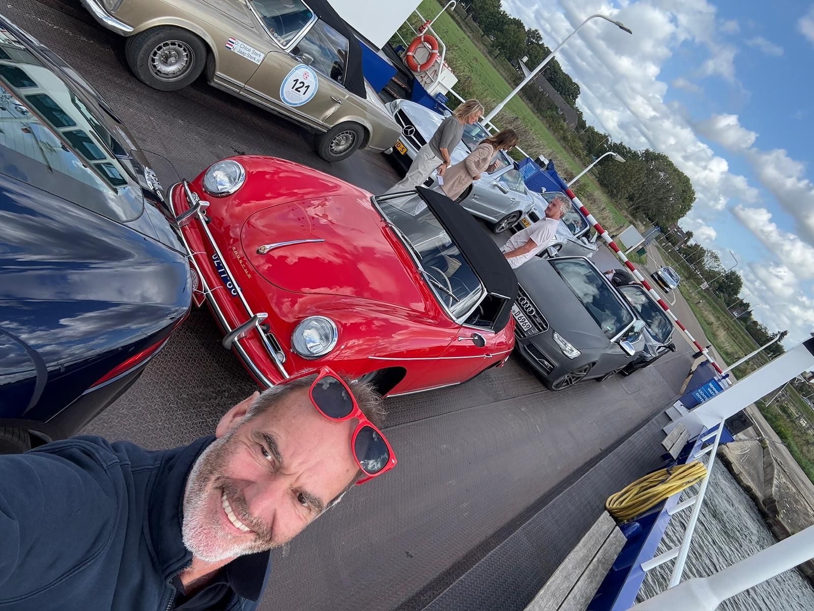 Een man poseert lachend voor een selfie met klassieke auto's op een veerboot, waarbij een rode cabriolet prominent in beeld is.