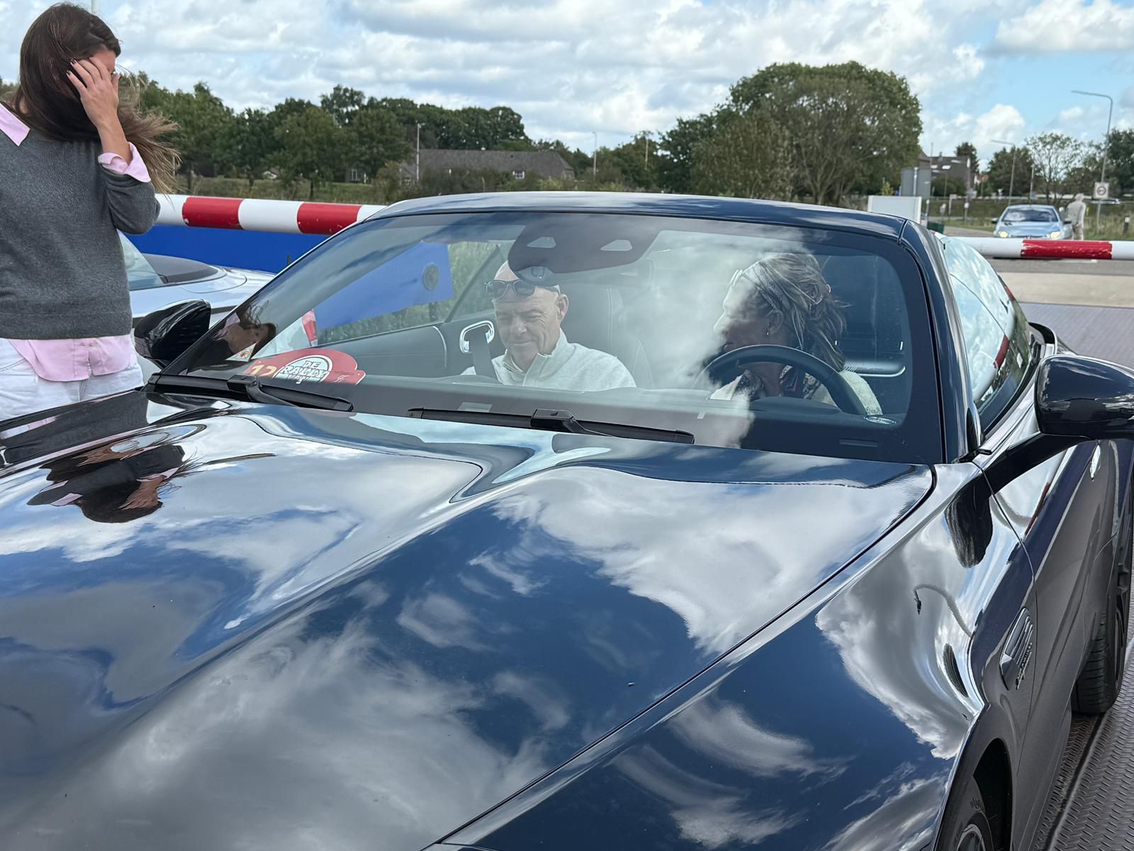 Zwarte auto met twee inzittenden, een vrouw ernaast met haar hand op haar hoofd, blauwe lucht.