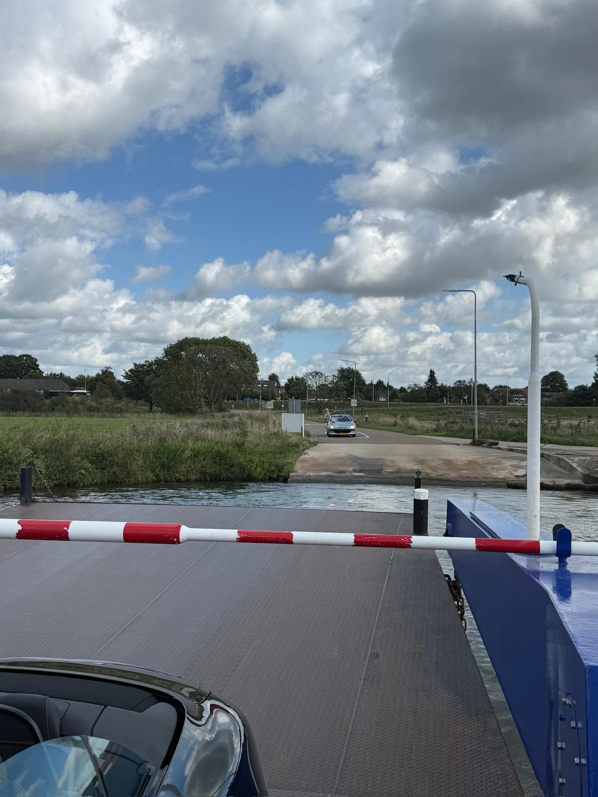 Een auto op een veerboot nadert een helling naar de wal, onder een bewolkte blauwe hemel. Op de voorgrond is een rood-wit gestreepte afscheiding te zien.