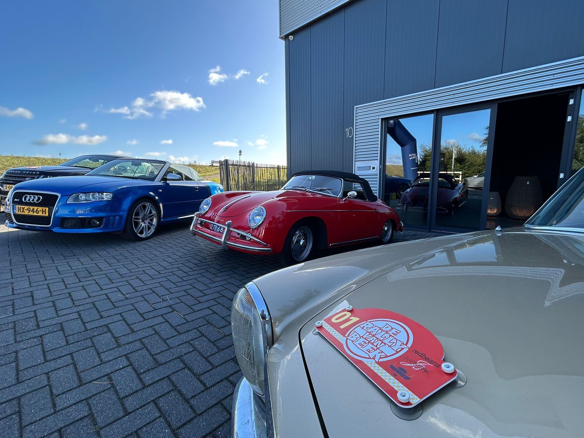 Op een zonnige dag stonden er diverse auto's, waaronder een rode Porsche en een blauwe cabriolet, geparkeerd voor een gebouw.