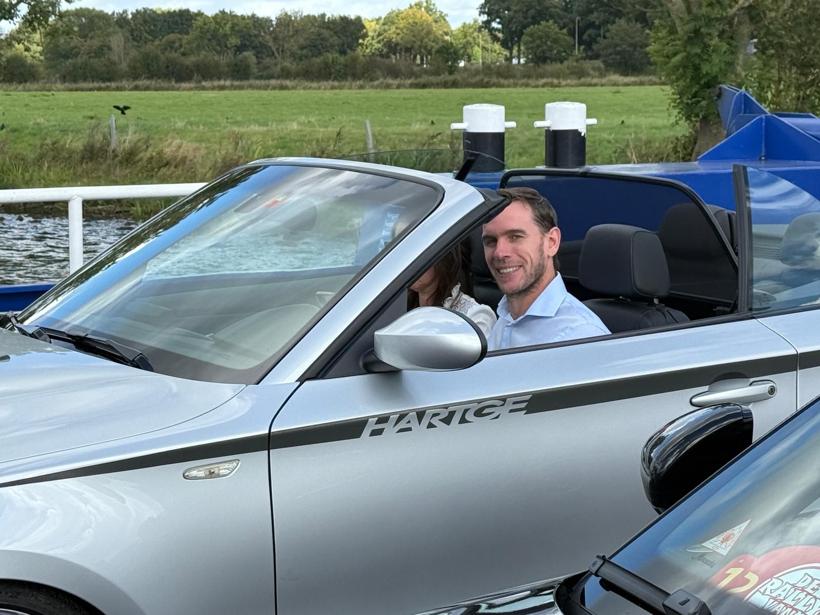 Een man in een zilverkleurige cabriolet glimlacht terwijl hij over een veerboot rijdt; water en groen op de achtergrond.