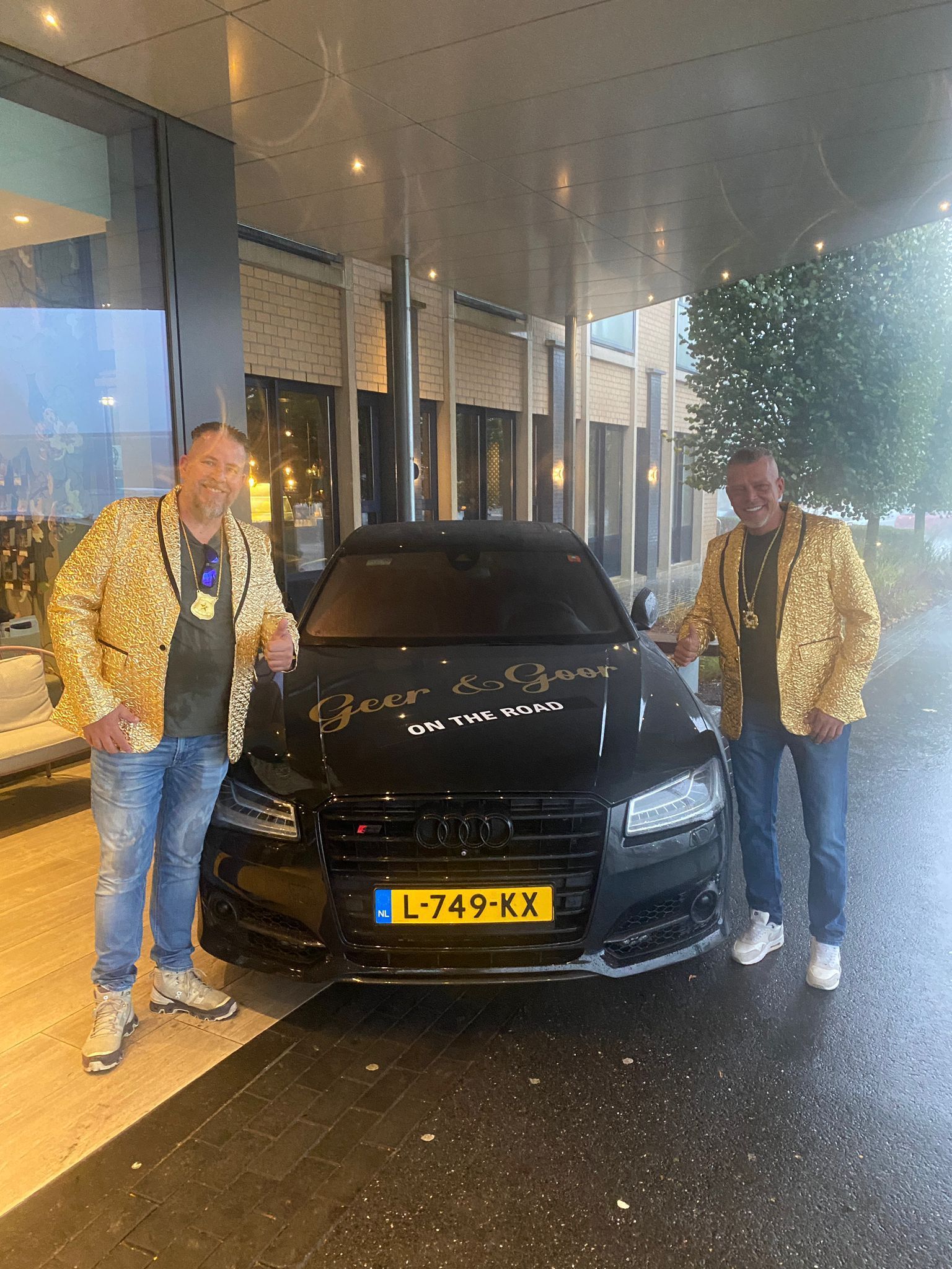 Twee mannen in gouden jasjes met pailletten poseren bij een zwarte Audi. Op de voorruit van de auto staat een boodschap.