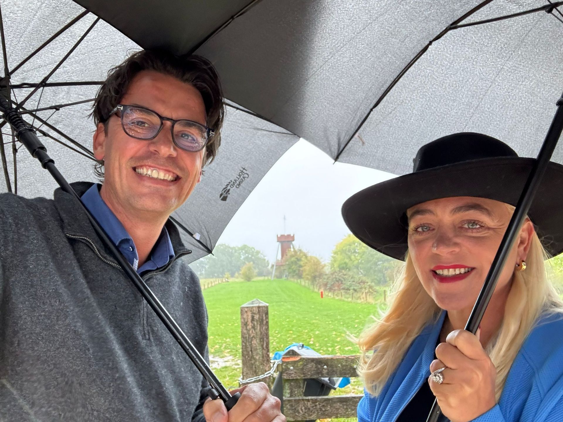 Twee mensen staan ​​lachend onder paraplu's in een regenachtige buitenomgeving, met een toren in de verte.