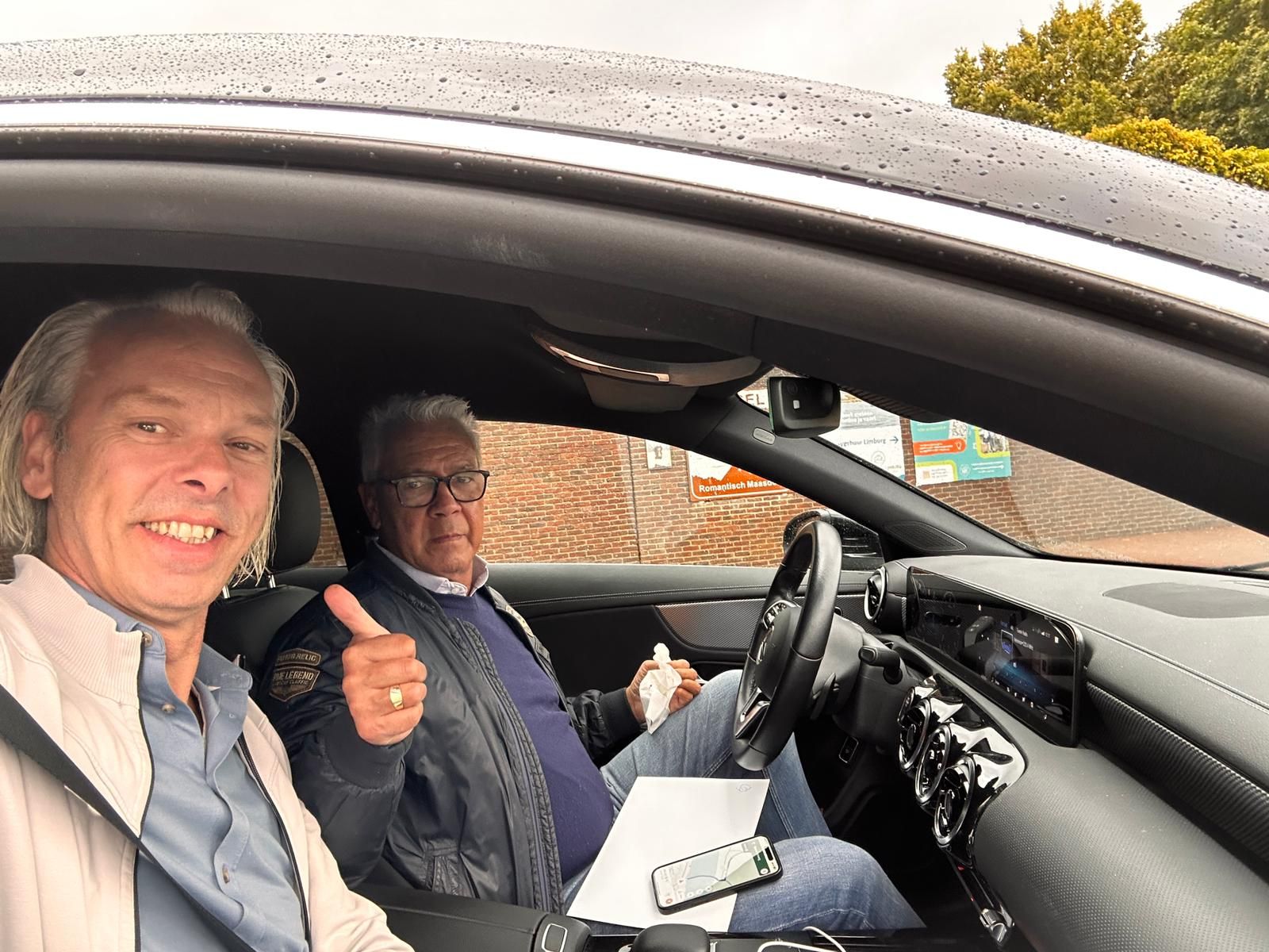 Twee mannen in een auto. De ene lacht en de andere steekt zijn duim omhoog.