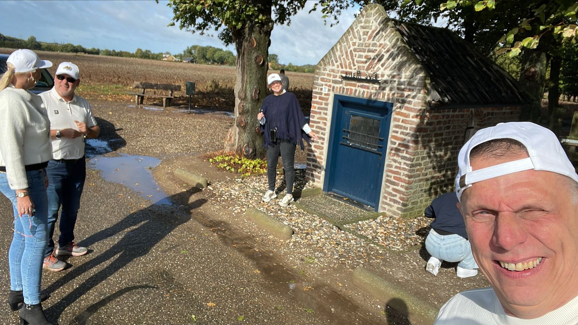 Mensen poseren bij een klein bakstenen gebouw met een blauwe deur, op een zonnige dag.