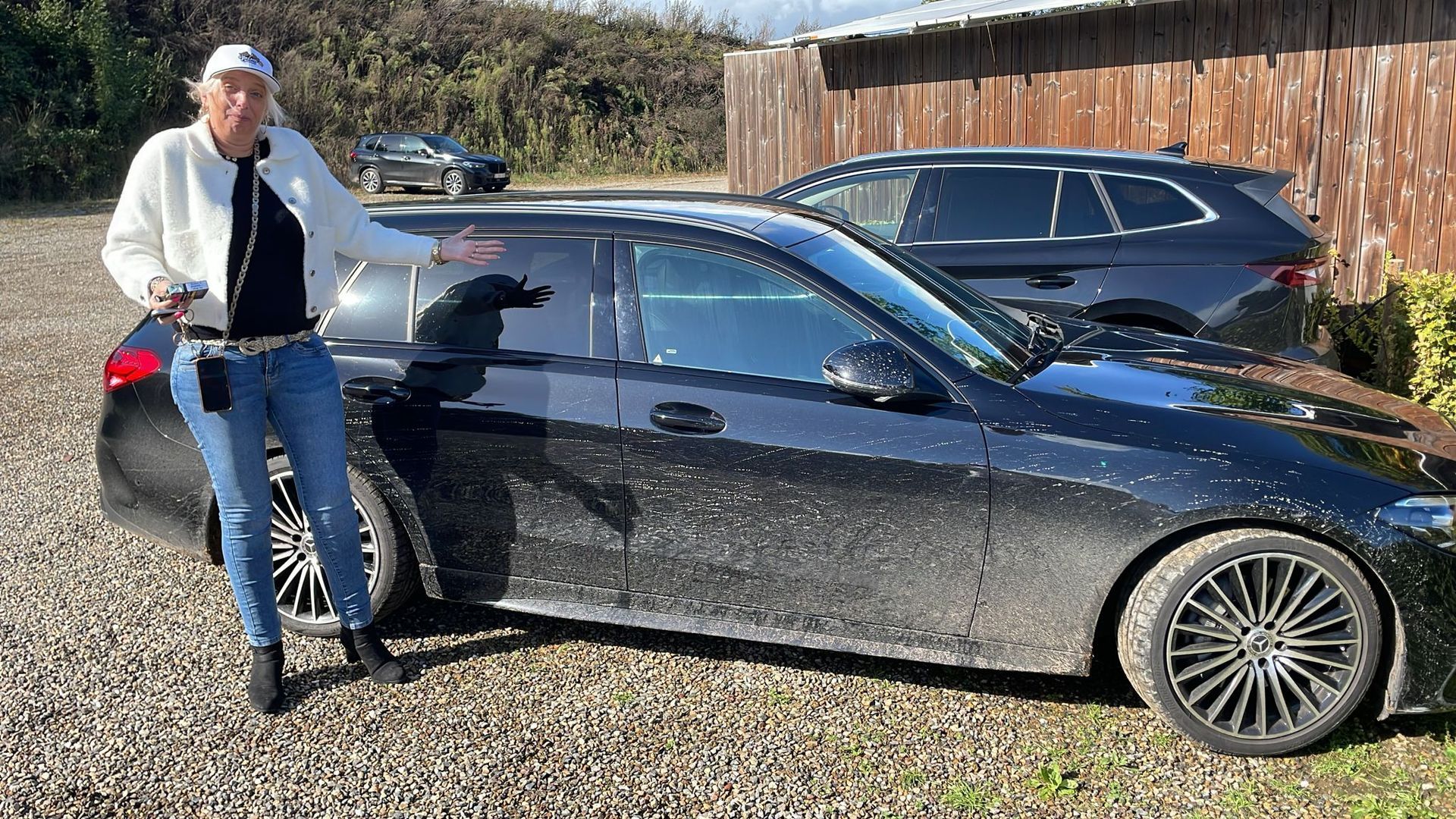 Een persoon staat naast een zwarte, met vuil bedekte auto en gebaart ernaar. Op de achtergrond is nog een auto te zien.