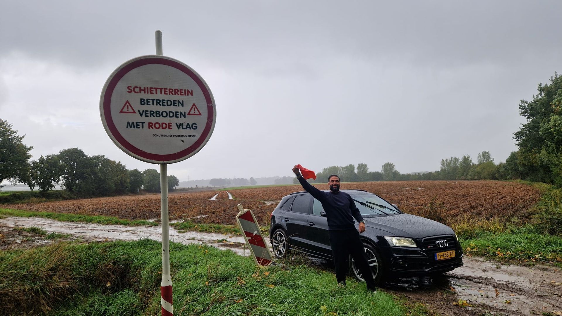 Man voor een auto bij een verkeersbord, rode vlag gehesen in een regenachtig veld. Op het bord staat 