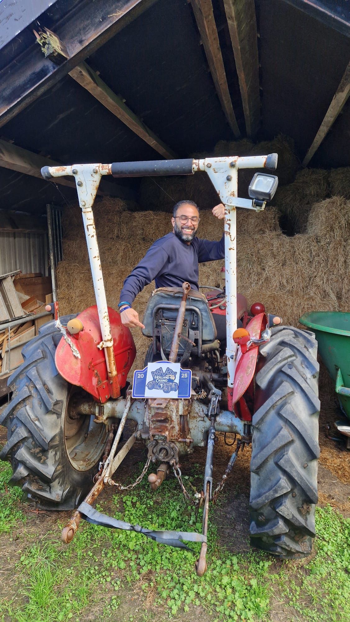 Een man zit op een rood-grijze tractor in een schuur. Hij lacht en houdt het tractorframe vast.