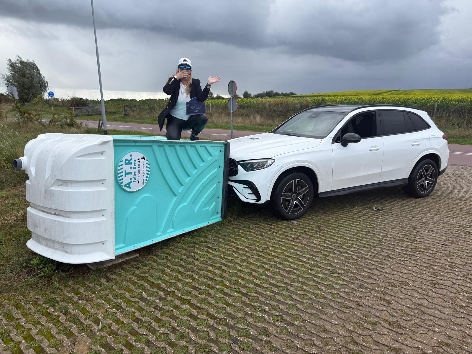 Een vrouw gebaart vanuit een mobiel toilet dat aan een witte SUV is gekoppeld. Bewolkte buitenomgeving.