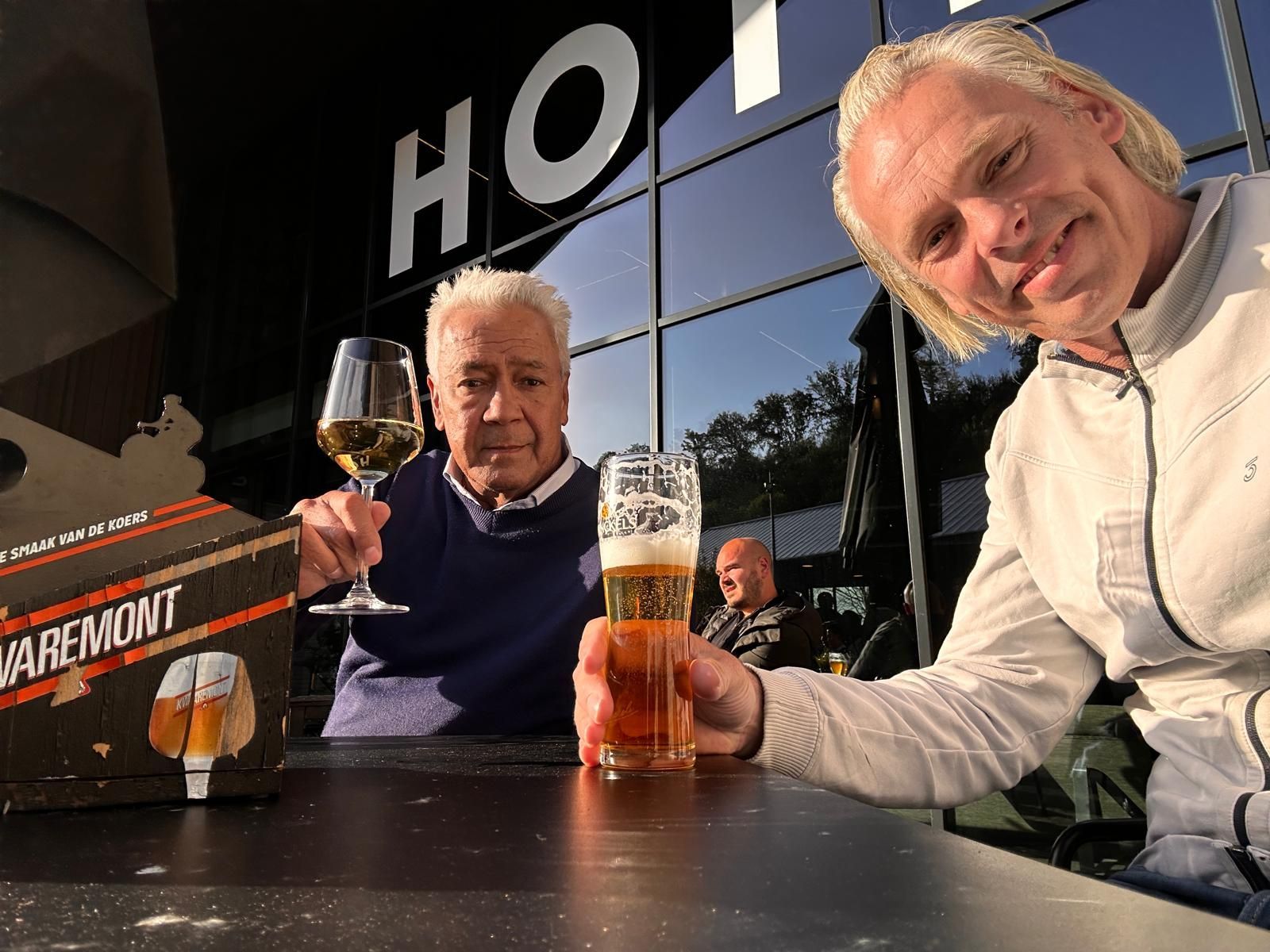 Twee mannen zitten aan een tafel en proosten met drankjes voor een gebouw met het opschrift 