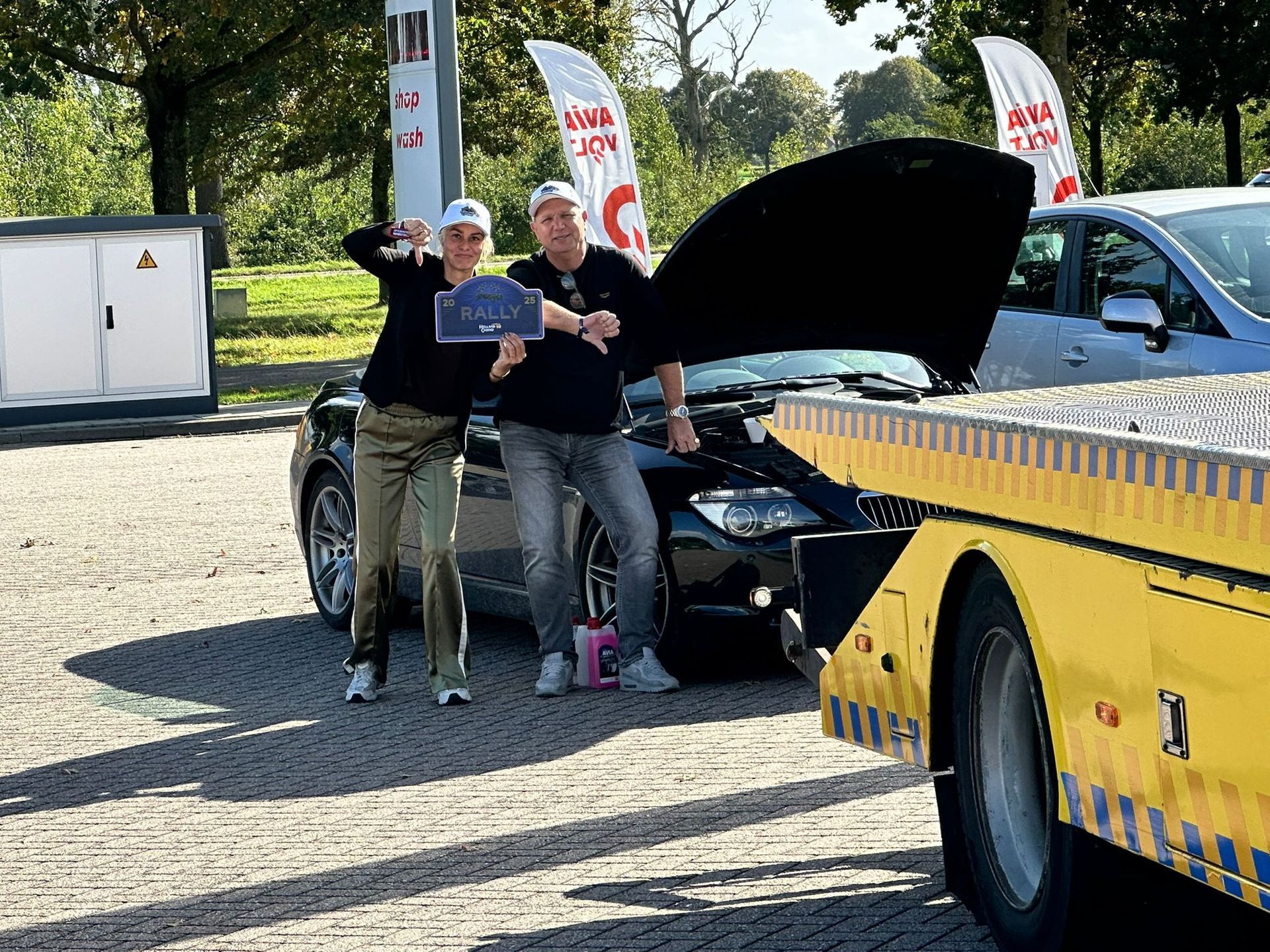 Twee mensen poseren naast een auto met open motorkap, voor een sleepwagen.
