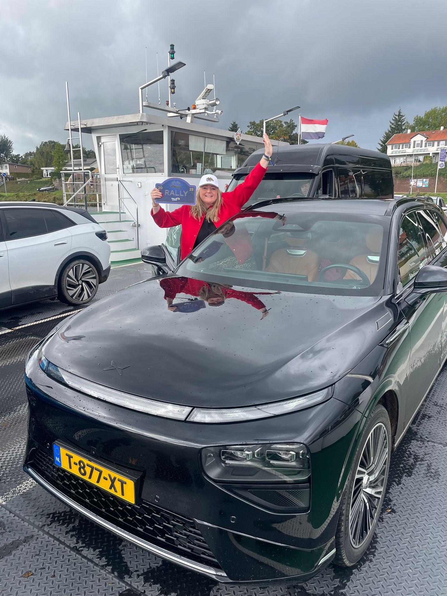 Vrouw in rode jas viert feest op een zwarte auto op een veerboot; Nederlandse vlag op de achtergrond.