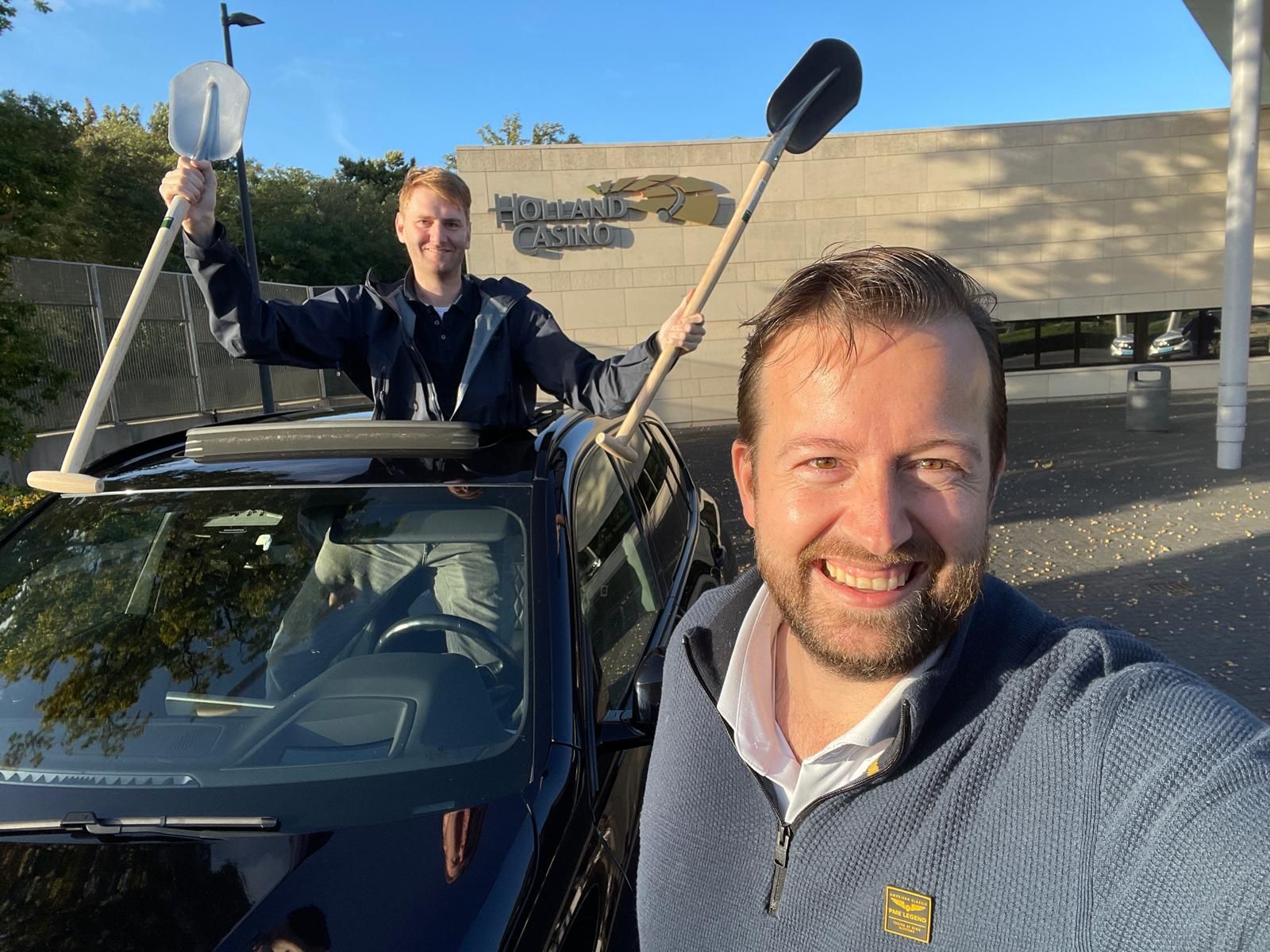 Twee mannen met schoppen in hun handen staan ​​lachend naast een auto bij een gebouw.