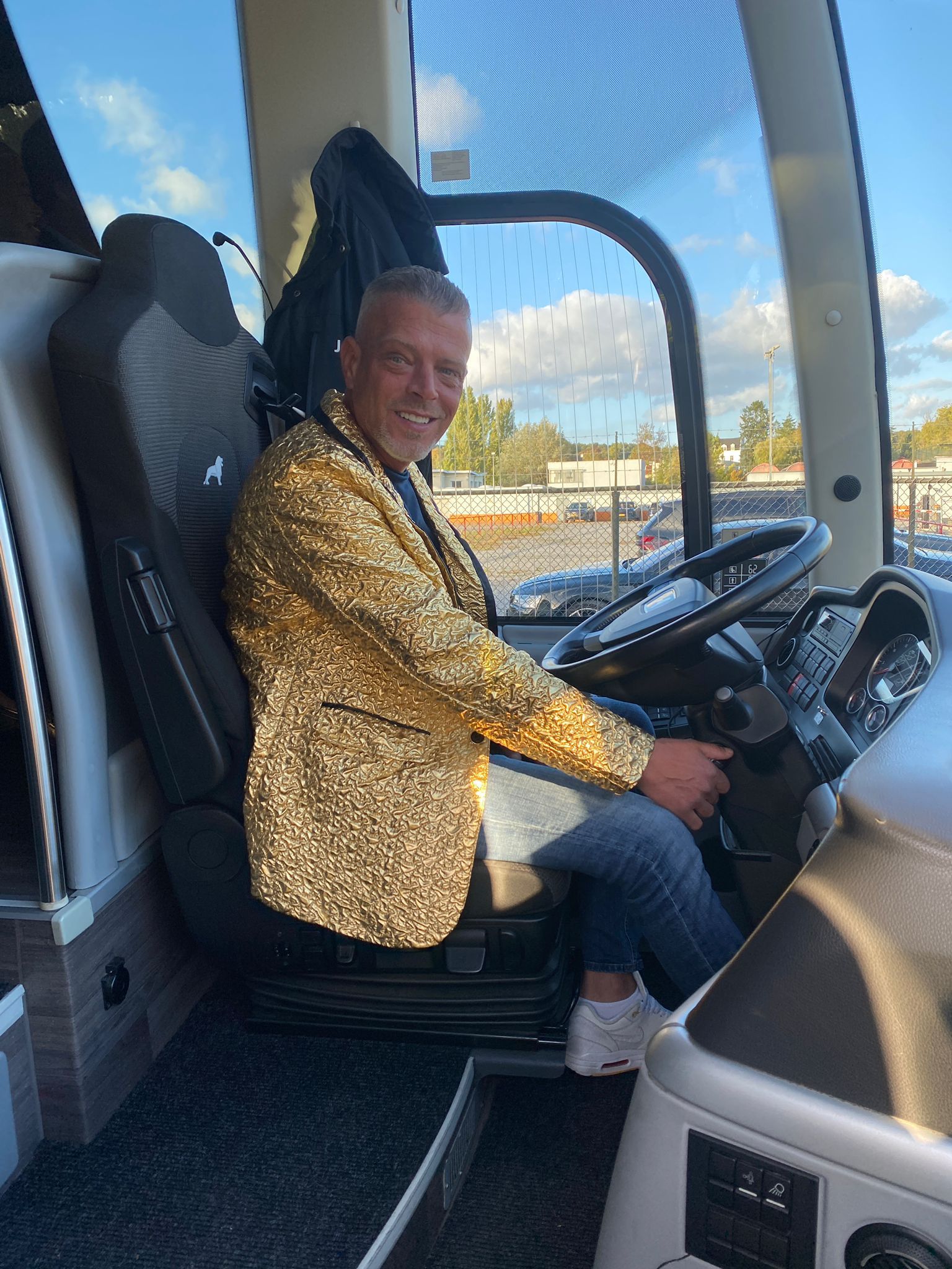 Man in gouden jasje met pailletten zit achter het stuur van een bus. Lachend, buiten.