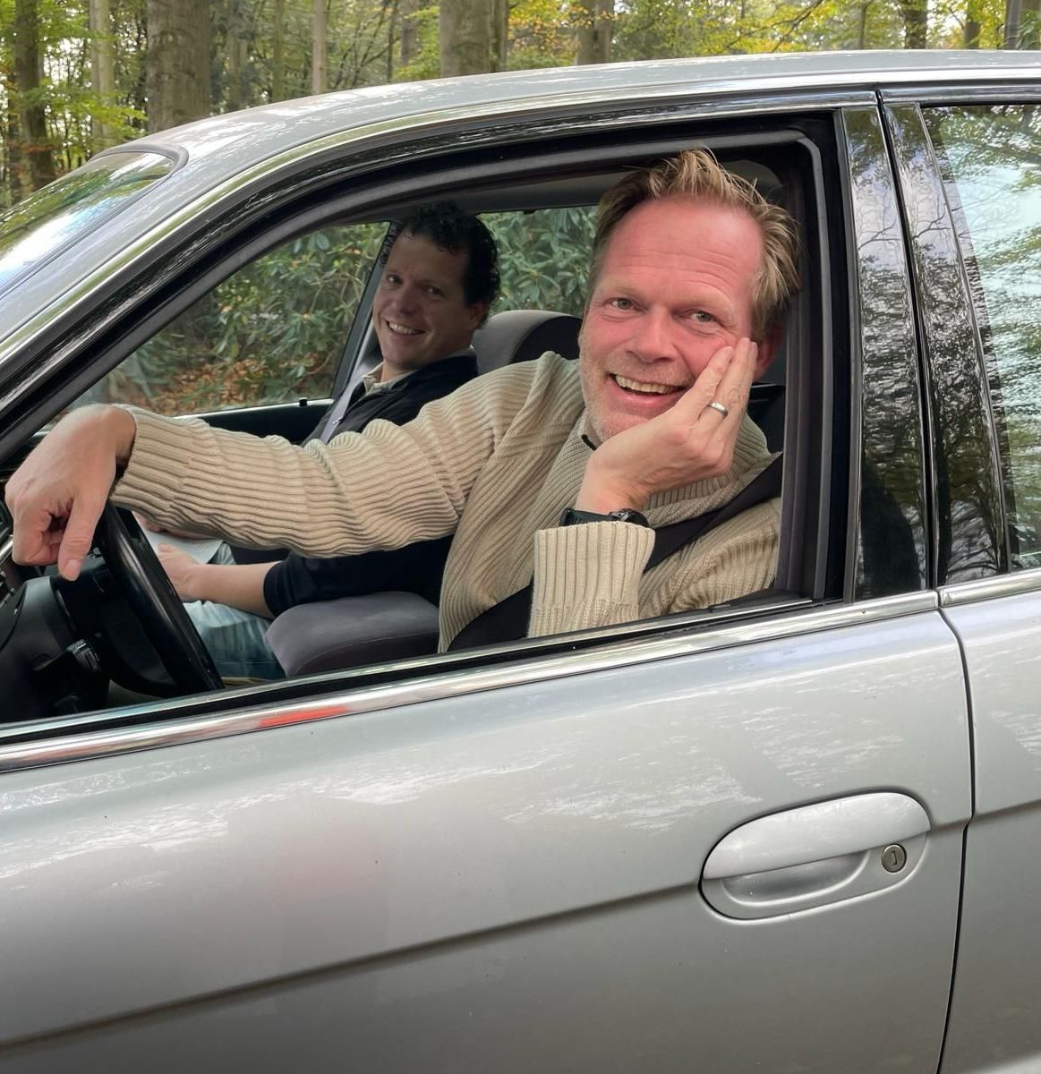 Twee lachende mannen in een zilverkleurige auto in een bosrijk gebied. De ene bestuurt de auto, de andere zit op de passagiersstoel.